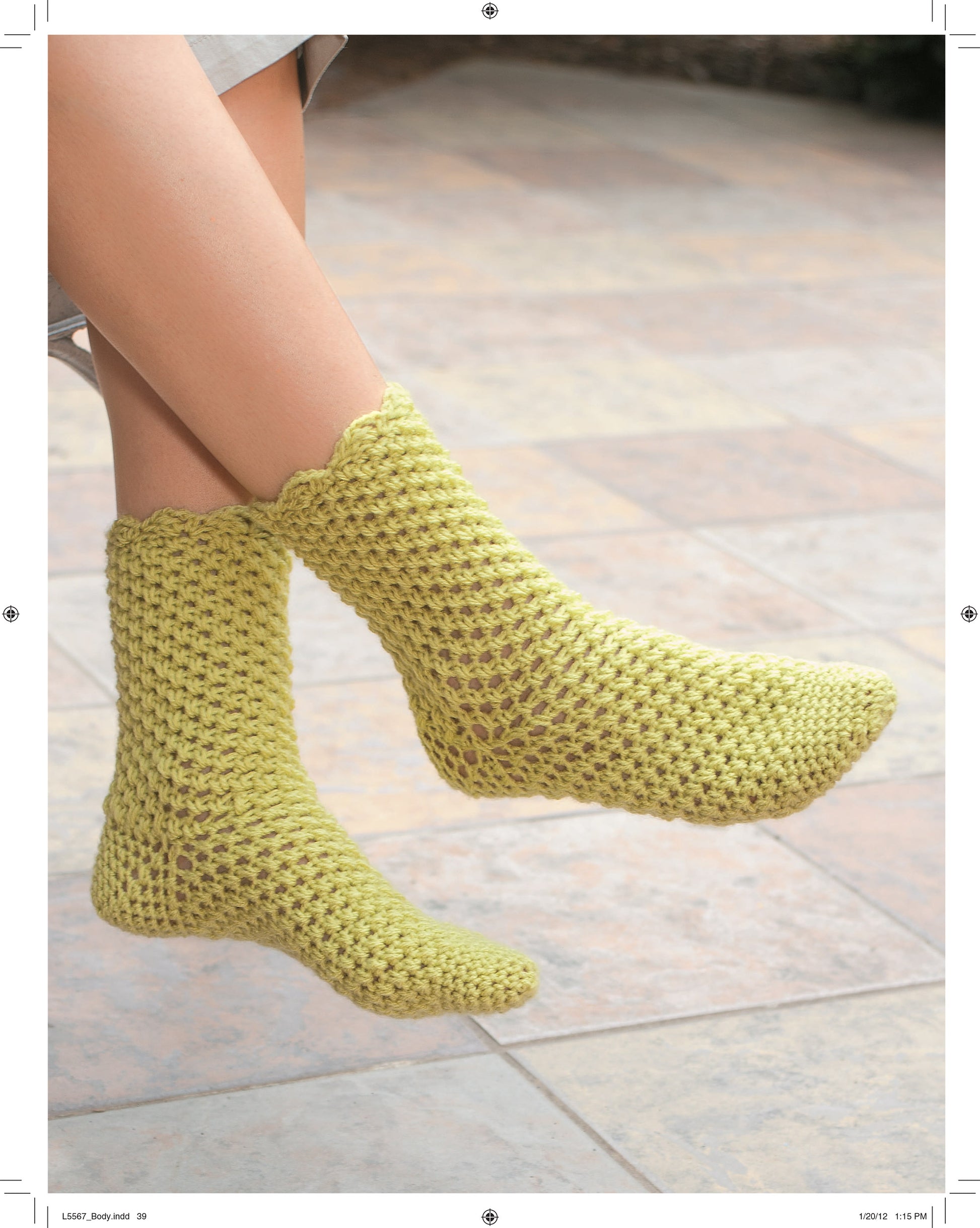 A person rests their feet on a tiled floor, showing only their lower legs and feet in light green hand-crocheted socks with a scalloped edge made using the “Socks for the Family” pattern by Leisure Arts.