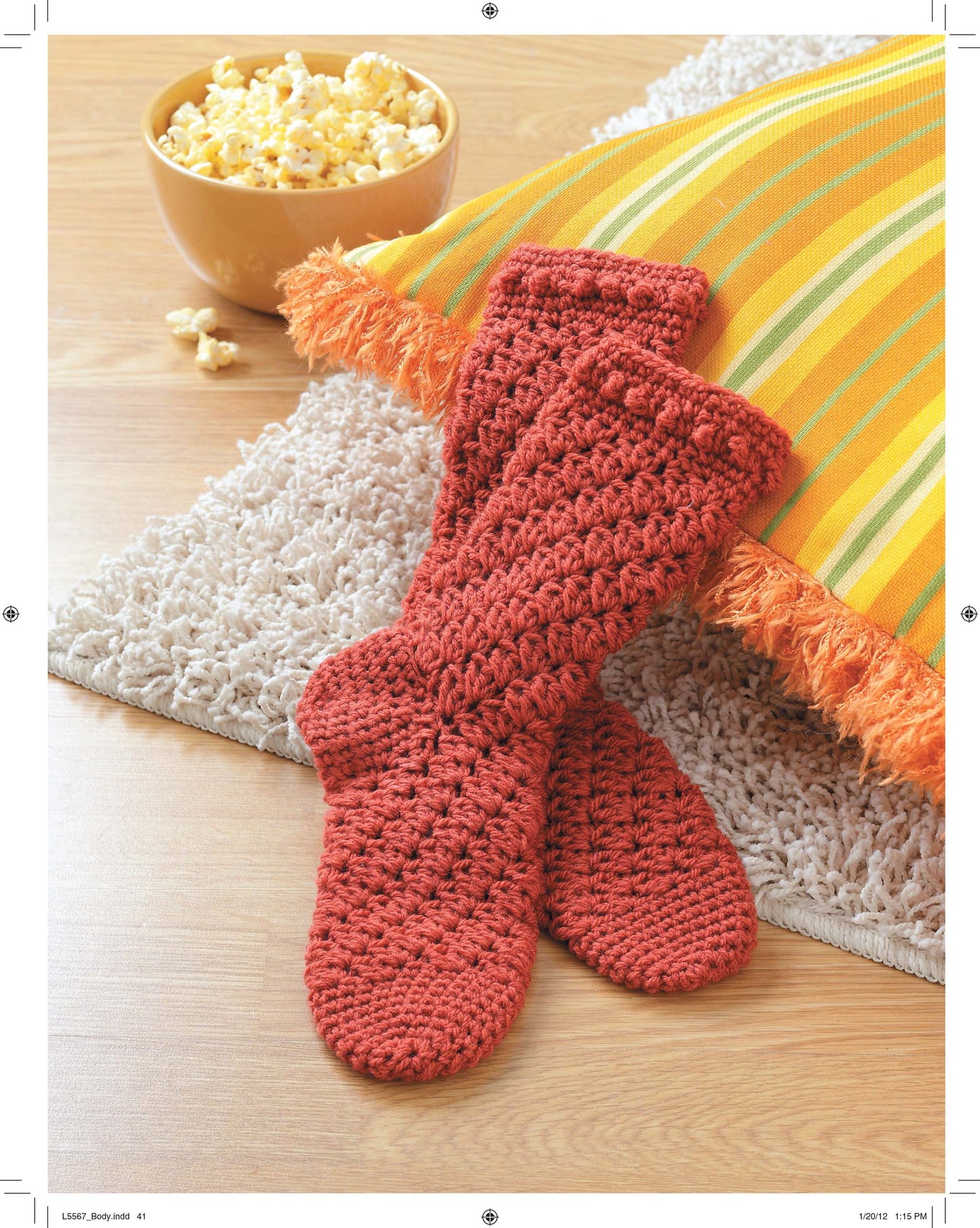 A pair of red crocheted socks made with the Socks for the Family pattern by Leisure Arts rests on a beige rug near a yellow and green striped fringe pillow, with a bowl of popcorn on the wooden floor.