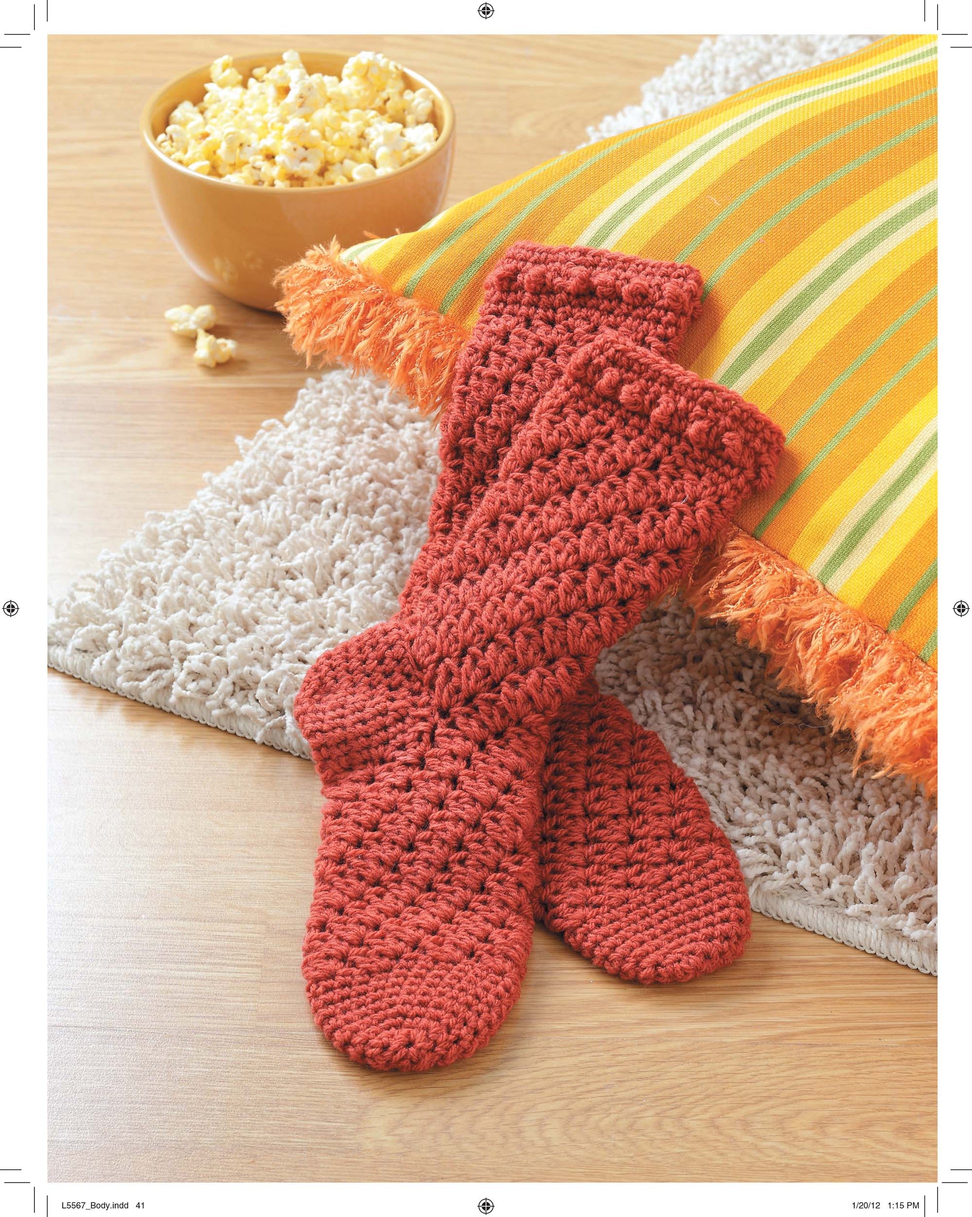 A pair of red crocheted socks made with the Socks for the Family pattern by Leisure Arts rests on a beige rug near a yellow and green striped fringe pillow, with a bowl of popcorn on the wooden floor.