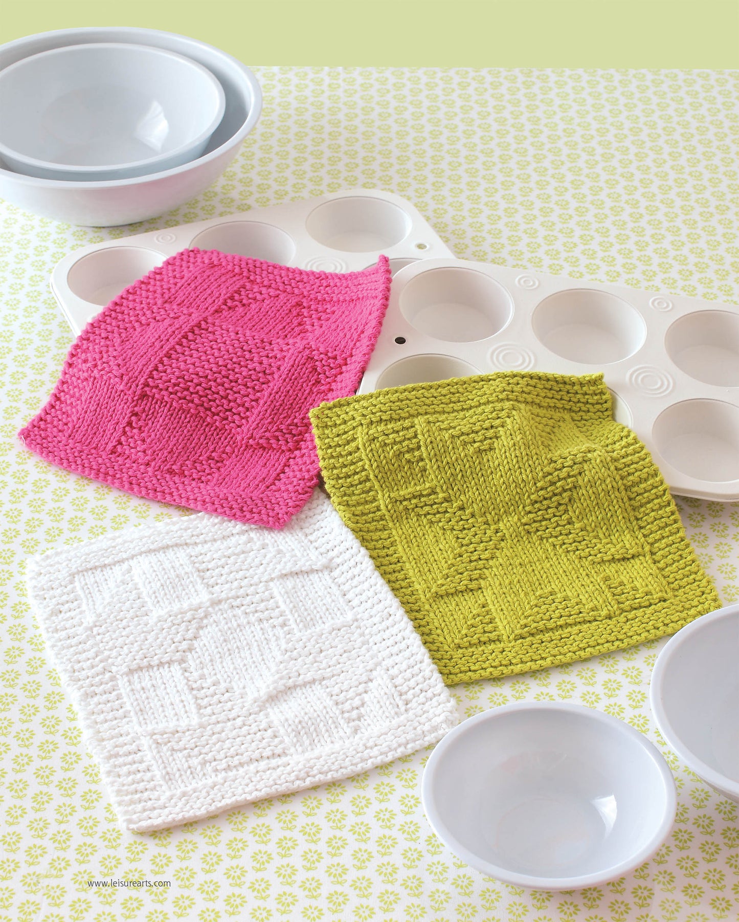 Leisure Arts Knit Dishcloths in pink, green, and white feature unique geometric knitting patterns. Set of three sits on a patterned surface beside white mixing bowls and a muffin tray, combining style with function.