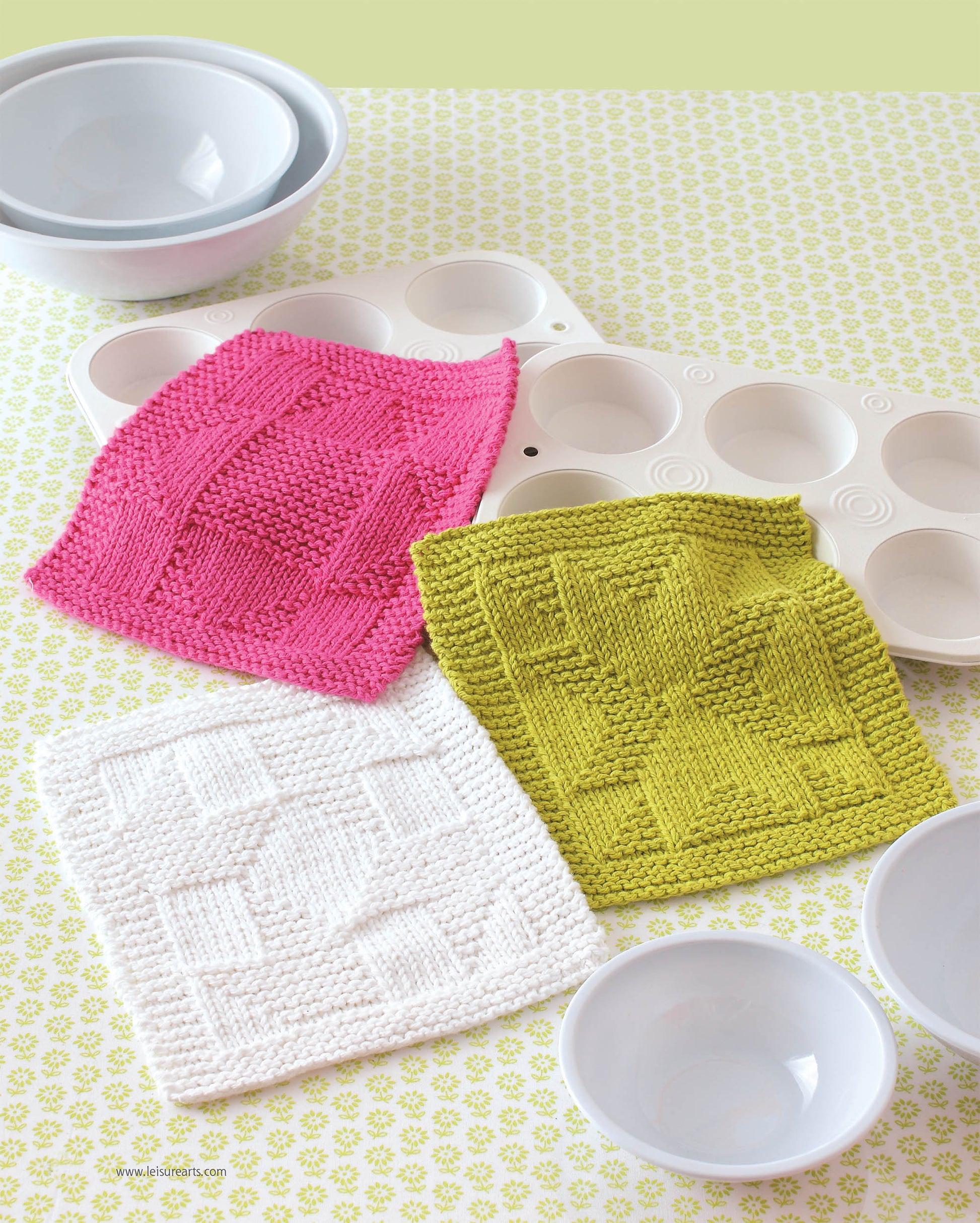 Leisure Arts Knit Dishcloths in pink, green, and white feature unique geometric knitting patterns. Set of three sits on a patterned surface beside white mixing bowls and a muffin tray, combining style with function.