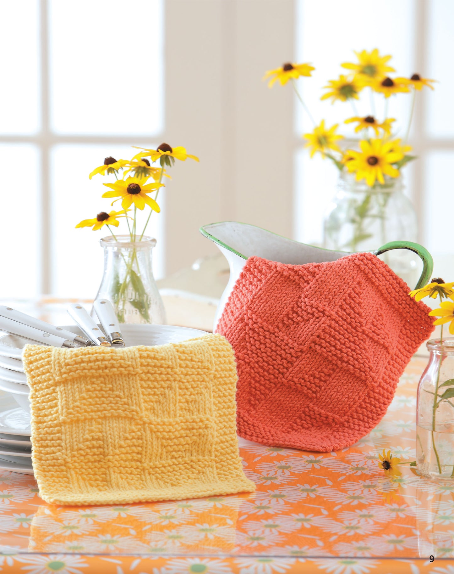 A yellow Leisure Arts Knit Dishcloth and a coral knitted pitcher cover rest on an orange floral tablecloth, with plates, utensils, and vases of yellow flowers nearby as sunlight streams through the window.