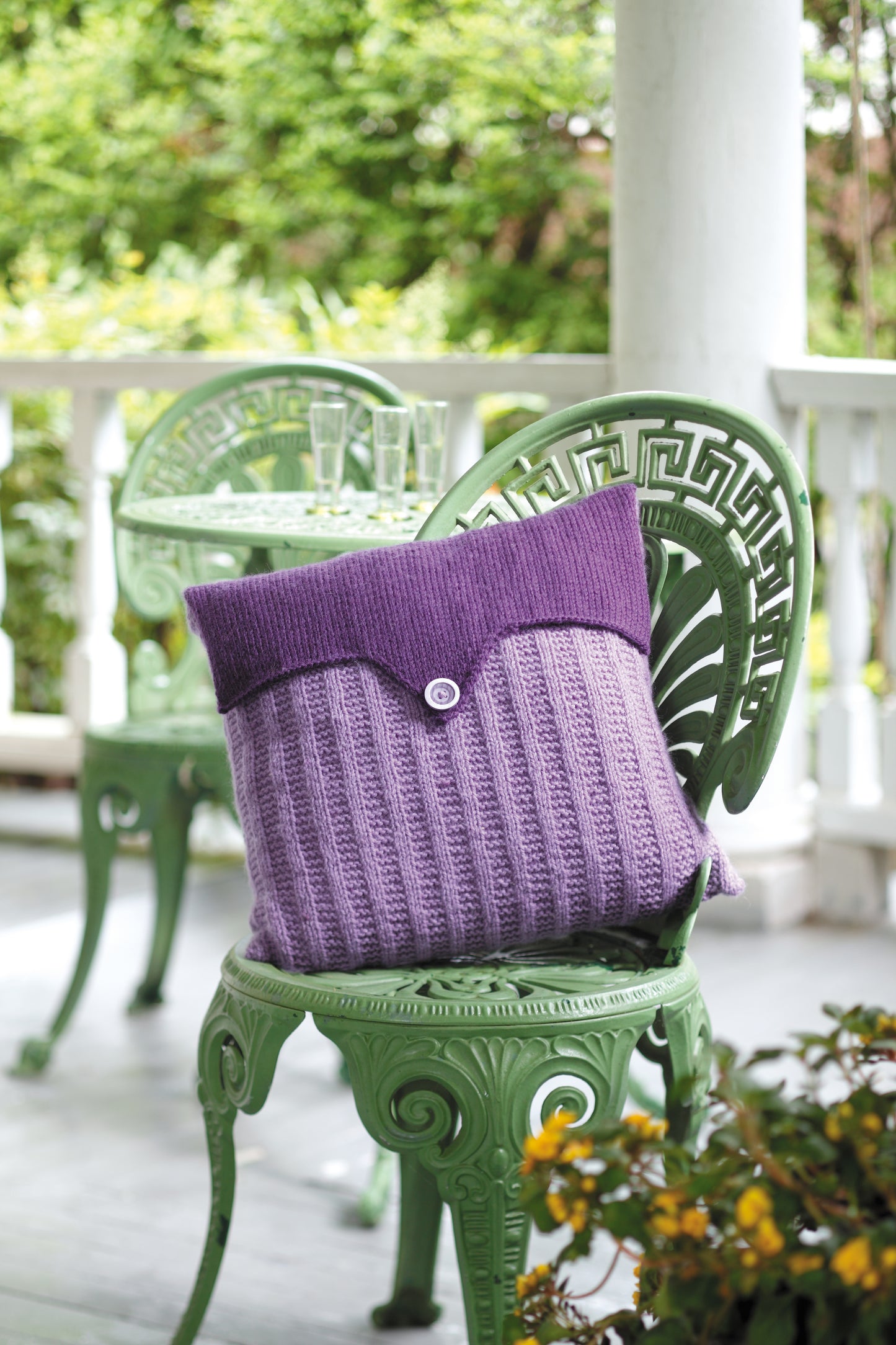 A purple knit pillow, inspired by Leisure Arts’ "The Envelope, Please!—Ten Knit Designs" digital pattern, rests on an ornate green chair on a porch with greenery and two empty champagne glasses nearby.