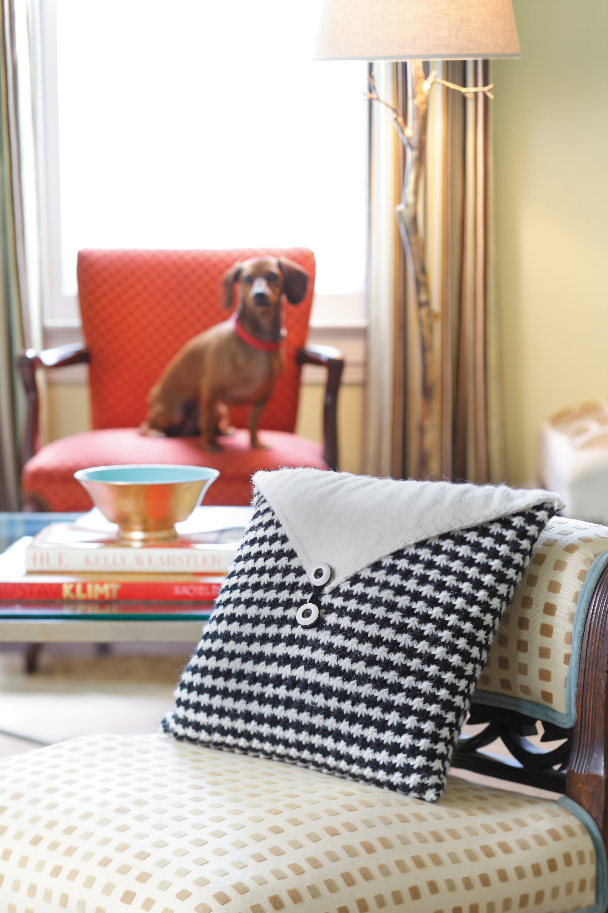 A living room features a black-and-white knit pillow on a cream armchair, made using The Envelope, Please!—Ten Knit Designs Digital Download by Leisure Arts; a small brown dog sits on a red chair near books and a lamp in the background.