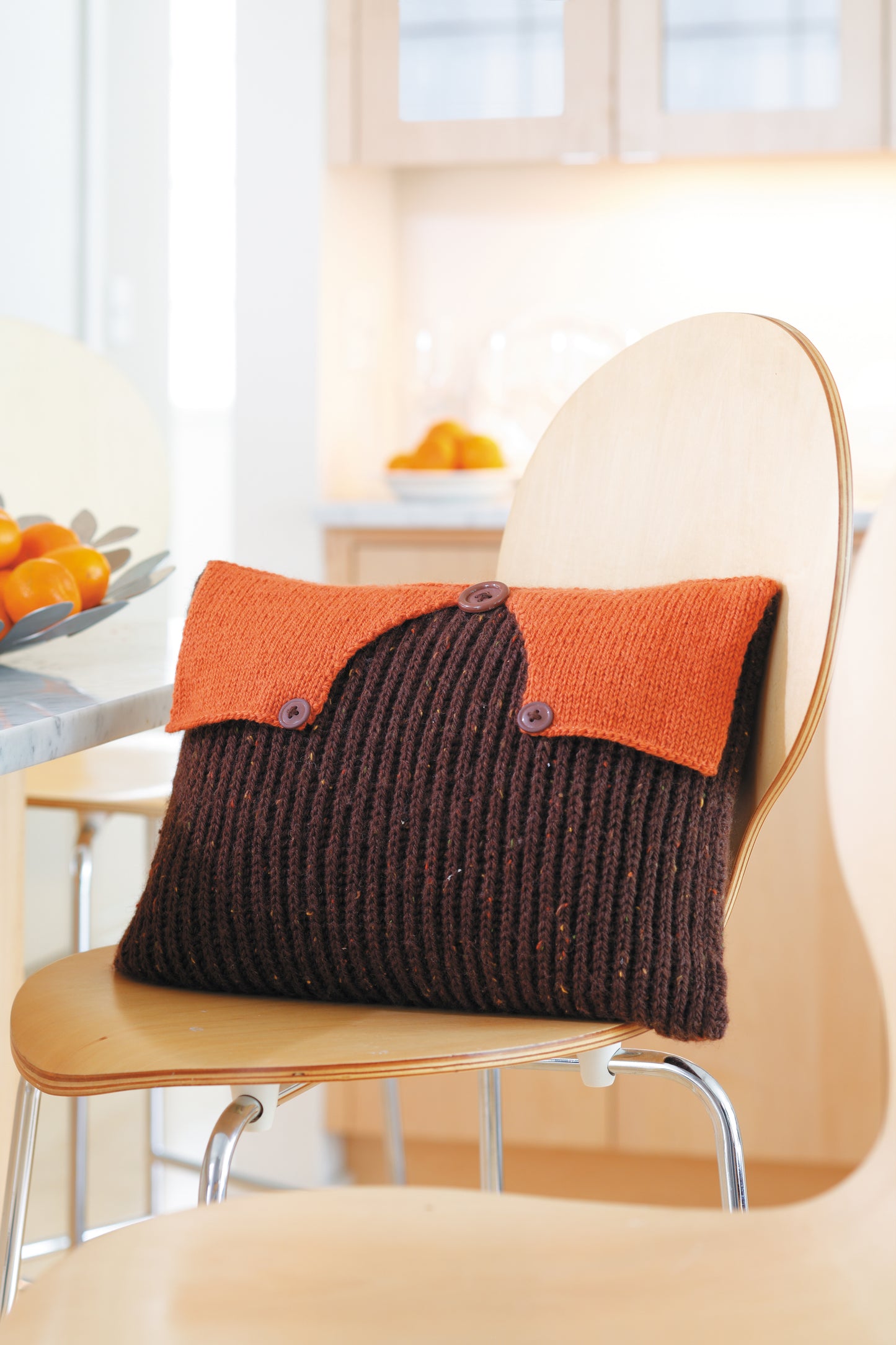 The Envelope, Please!—Ten Knit Designs by Leisure Arts features a worsted weight, ribbed brown pillow with an orange buttoned flap, displayed on a light wooden chair in a modern kitchen.