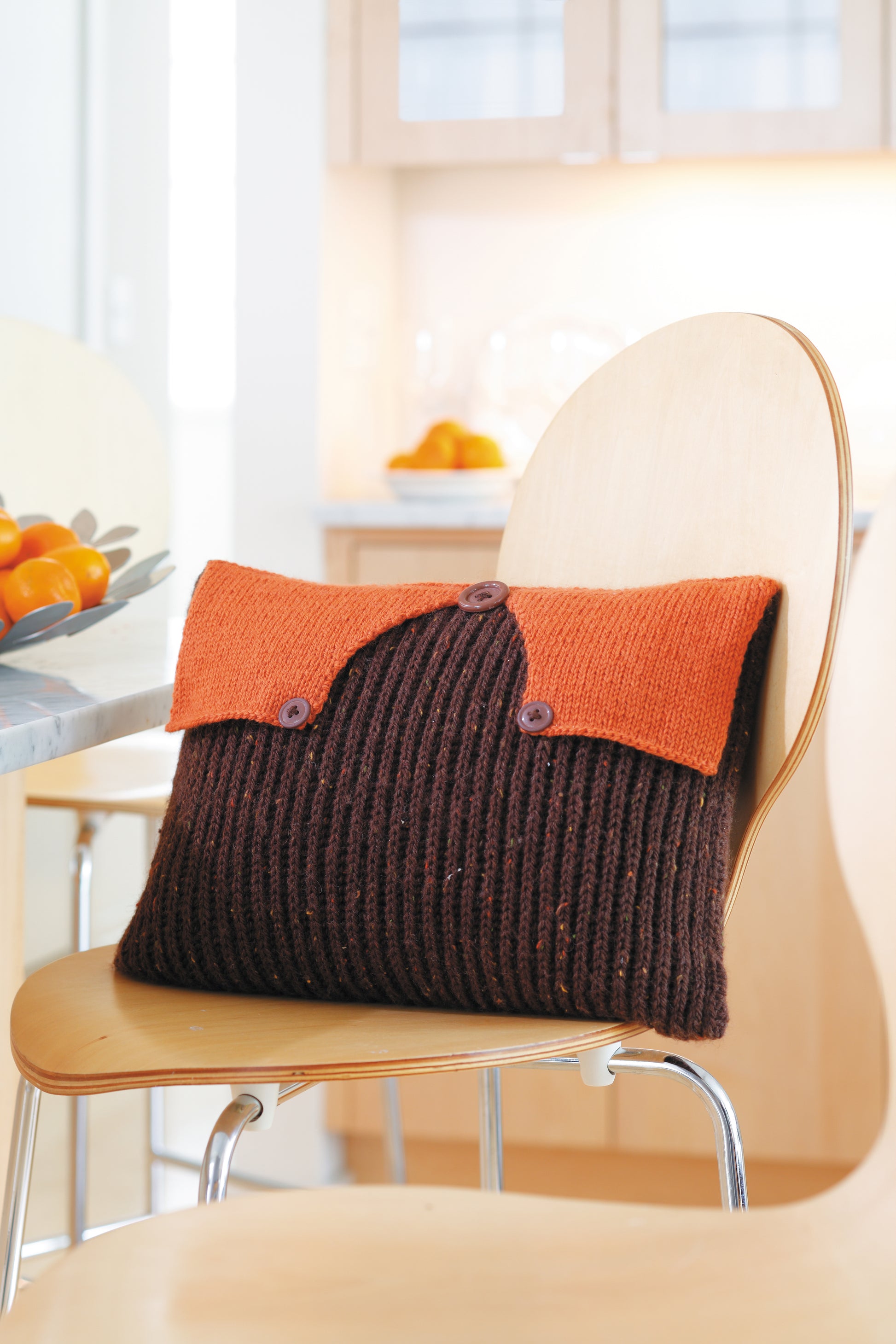The Envelope, Please!—Ten Knit Designs by Leisure Arts features a worsted weight, ribbed brown pillow with an orange buttoned flap, displayed on a light wooden chair in a modern kitchen.