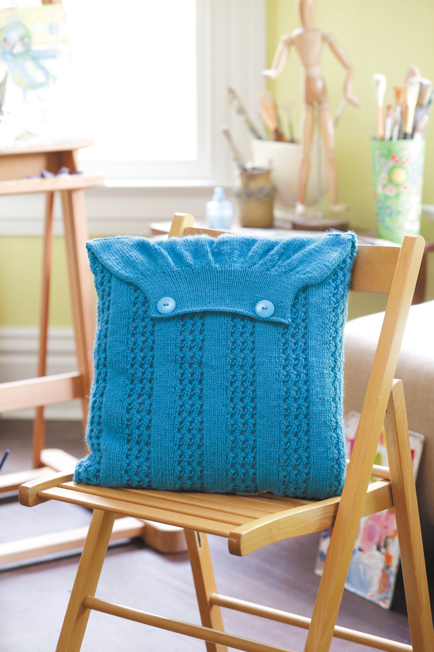 A wooden chair features a blue, cardigan-inspired pillow made from worsted yarn with buttons. Nearby, art supplies and a mannequin sit in a bright room. Design from The Envelope, Please!—Ten Knit Designs Digital Download by Leisure Arts.