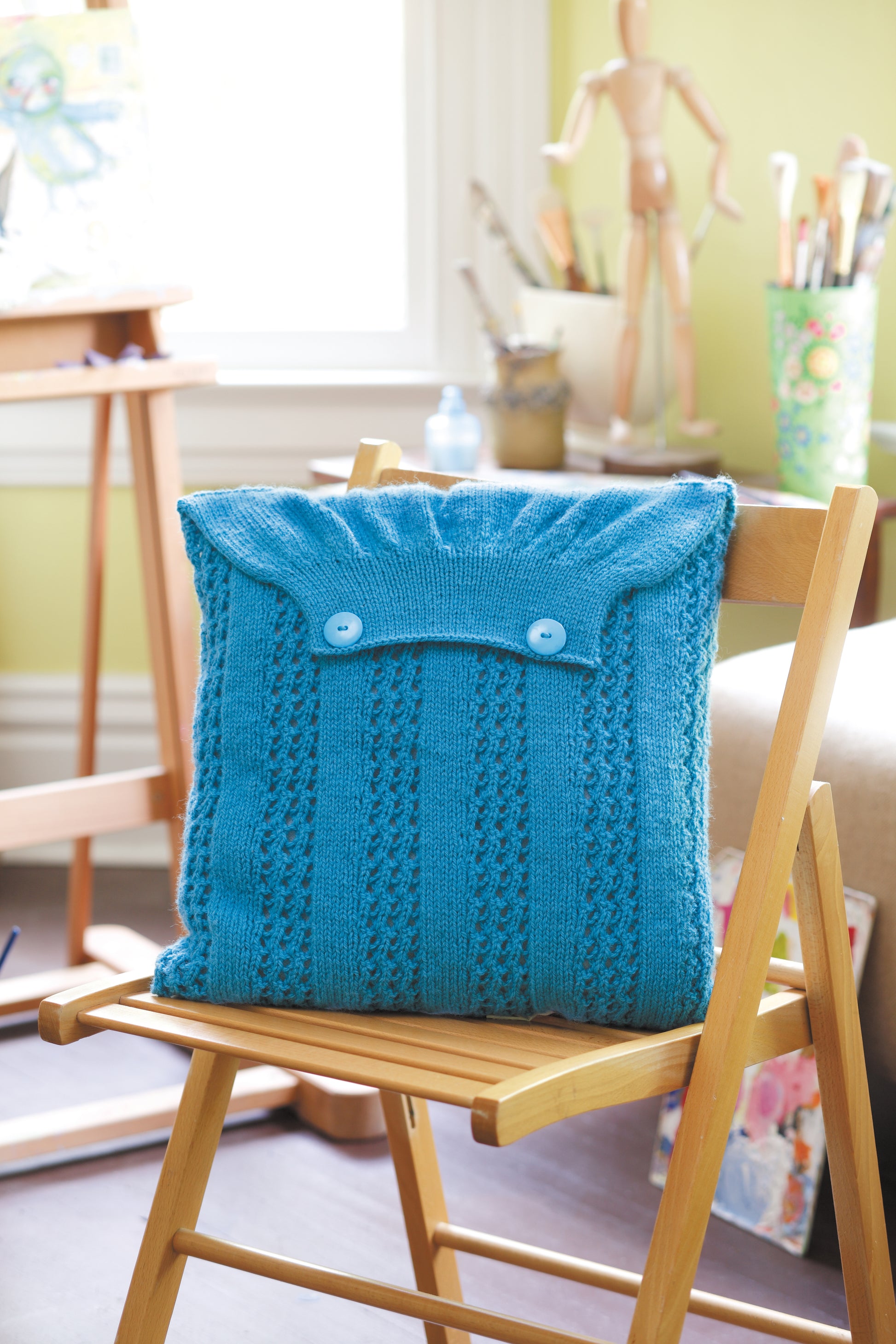 A wooden chair features a blue, cardigan-inspired pillow made from worsted yarn with buttons. Nearby, art supplies and a mannequin sit in a bright room. Design from The Envelope, Please!—Ten Knit Designs Digital Download by Leisure Arts.