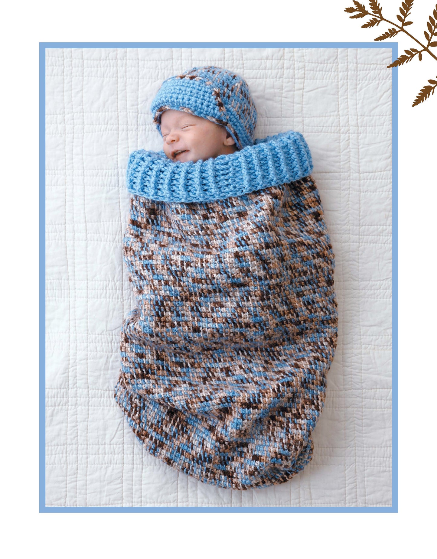 A newborn sleeps on a white quilt, wrapped in a Leisure Arts Crochet Dreamy Cocoons sack and cap in blue, brown, and white. A blue border frames the image with a brown leaf design at the top right.