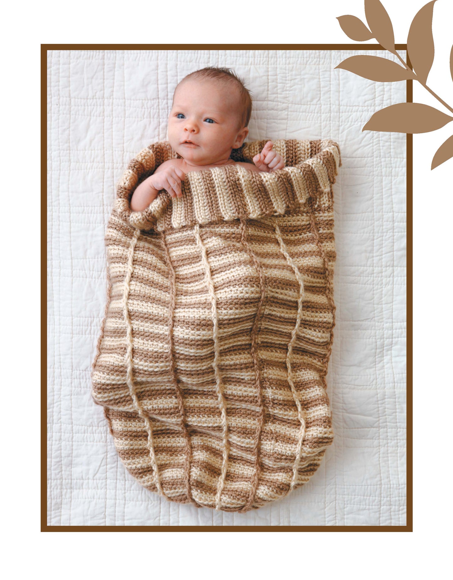 A baby lies on a white quilt, cozily wrapped in a Leisure Arts Crochet Dreamy Cocoons beige and brown striped cocoon. Brown leaves accent the top right border of the photo.