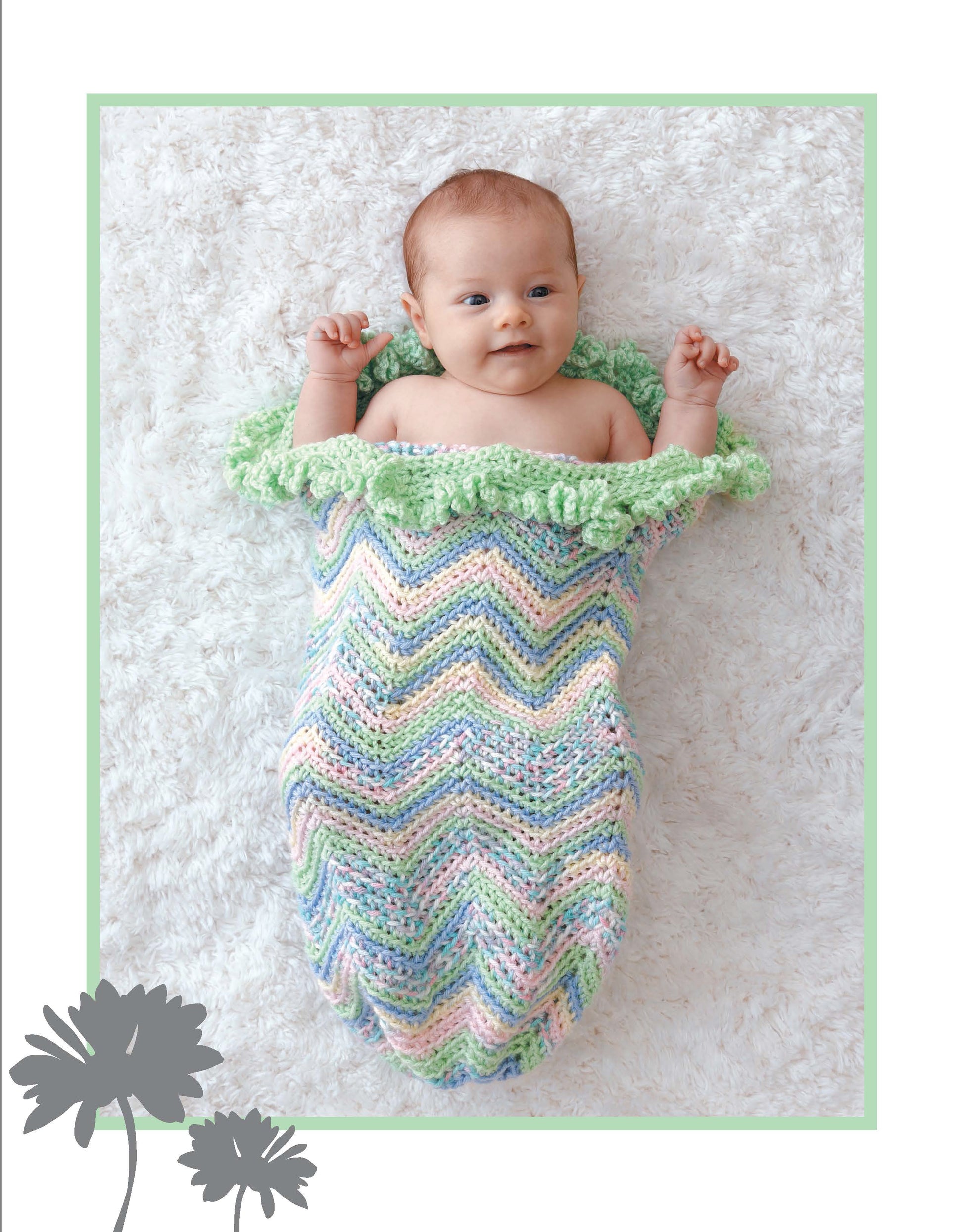A baby rests on a soft white rug, wrapped in the Leisure Arts Crochet Dreamy Cocoons—a pastel crochet cocoon with a ruffled green edge. Two gray flower silhouettes decorate the bottom left corner of the image.