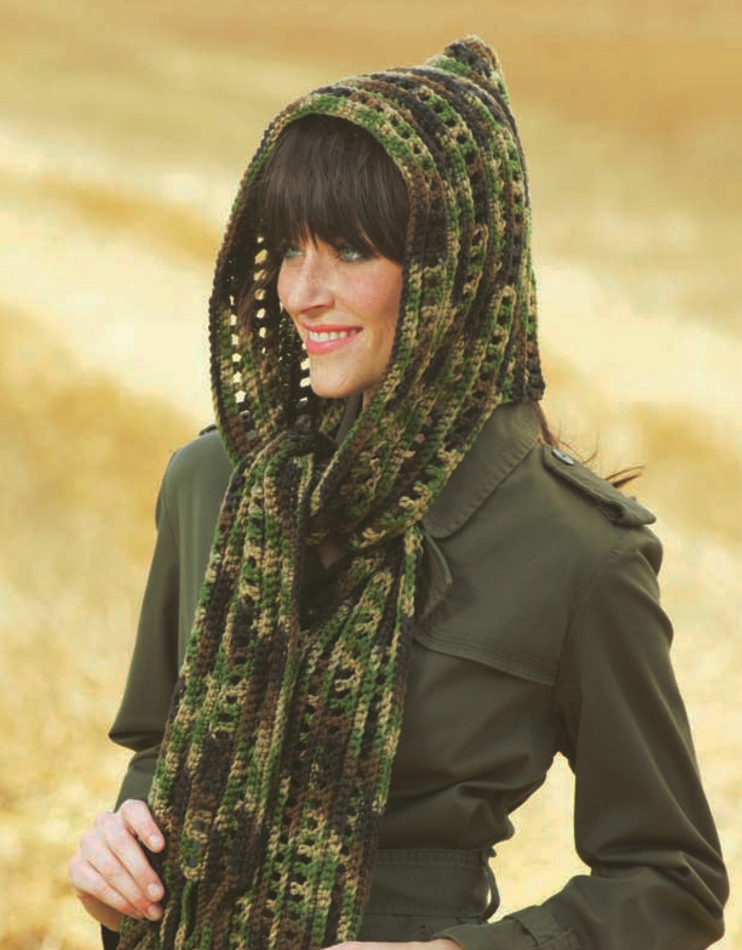 A woman stands outside, smiling in a blurred golden field, wearing a green, brown, and black hooded scarf made from the "Hooded Scarves Digital Download" crochet pattern by Leisure Arts—ideal for beginners.