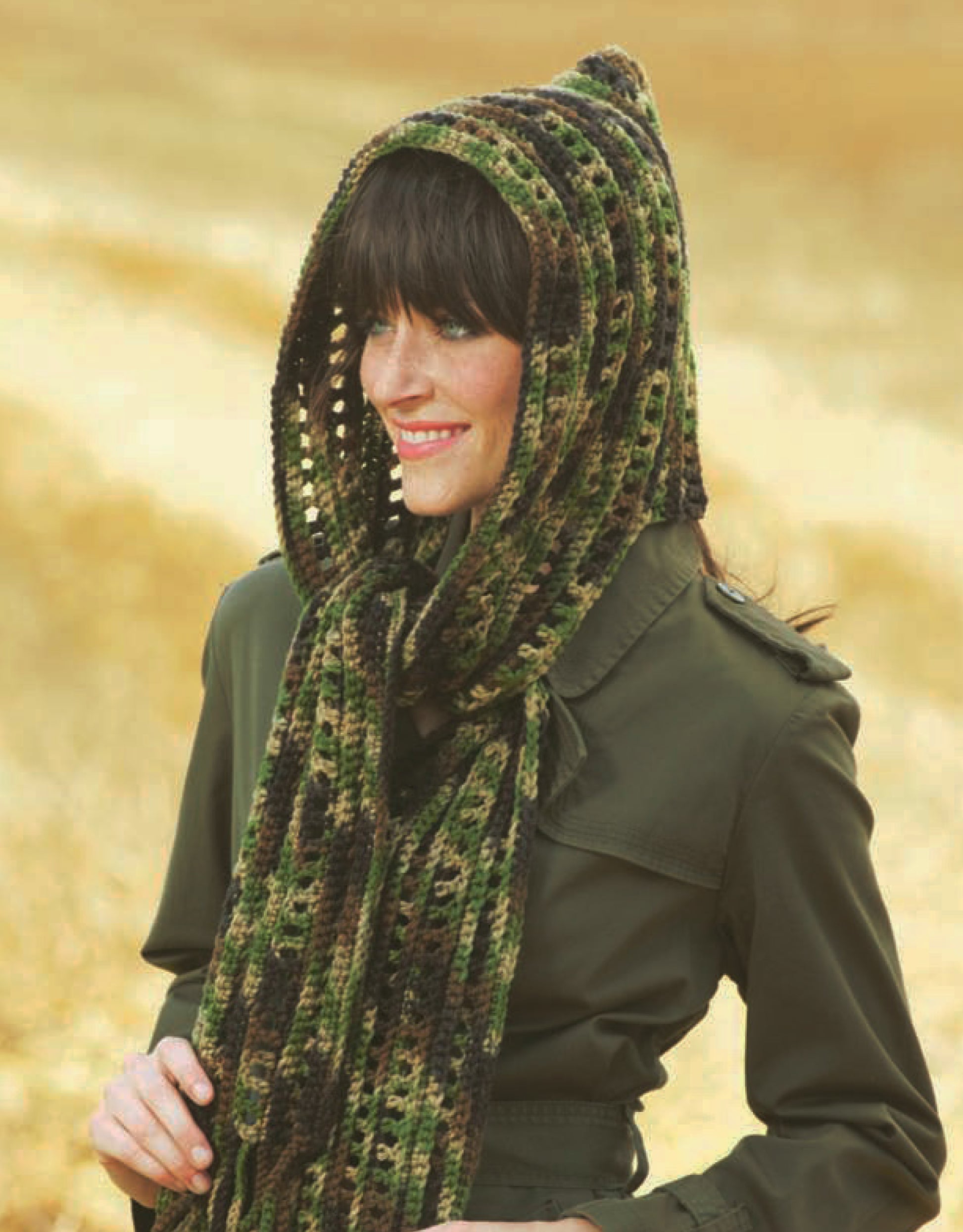 A woman stands outside, smiling in a blurred golden field, wearing a green, brown, and black hooded scarf made from the "Hooded Scarves Digital Download" crochet pattern by Leisure Arts—ideal for beginners.
