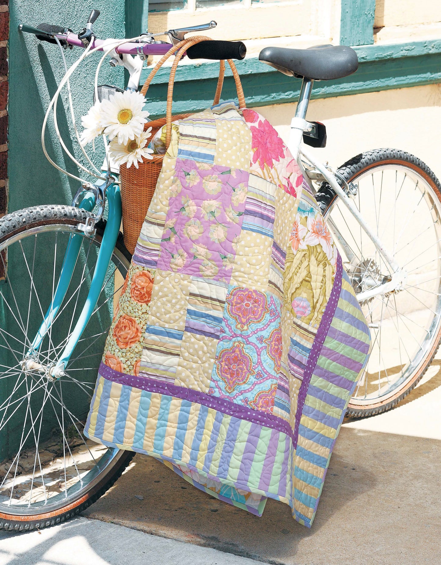 A white bicycle on the sidewalk features a wicker basket of white daisies and the colorful Leisure Arts "Fresh, Fast & Fun Quilts" draped over the handlebars and wheel, creating a bright, cheerful scene near a painted building.