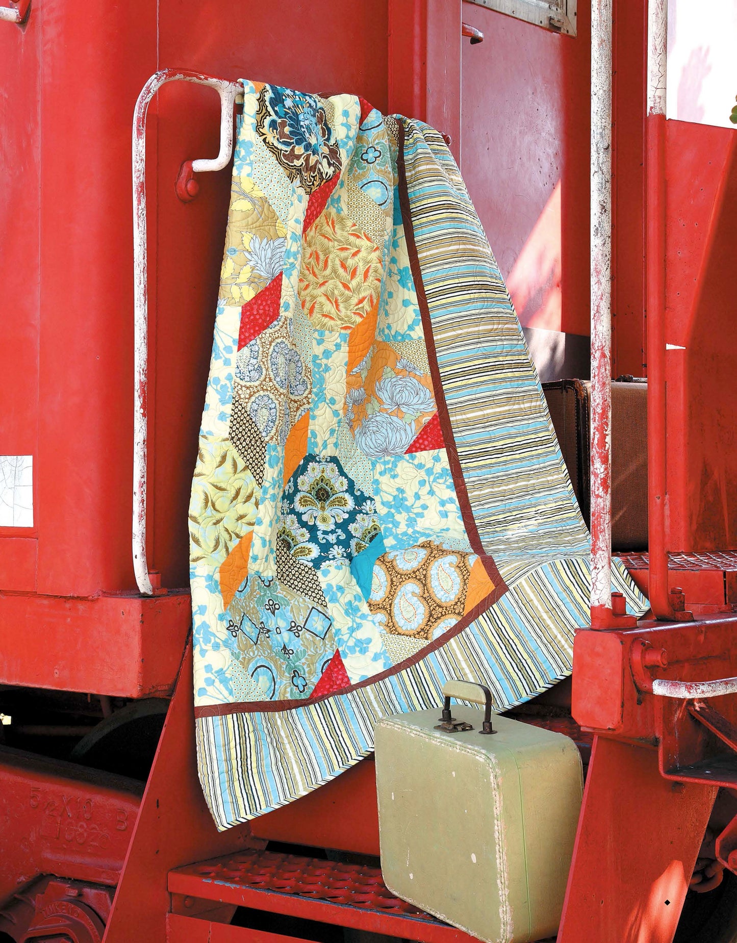 The "Fresh, Fast & Fun Quilts" by Leisure Arts features vibrant floral and geometric designs, displayed here draped over a bright red train car with a light green vintage suitcase on the step below.