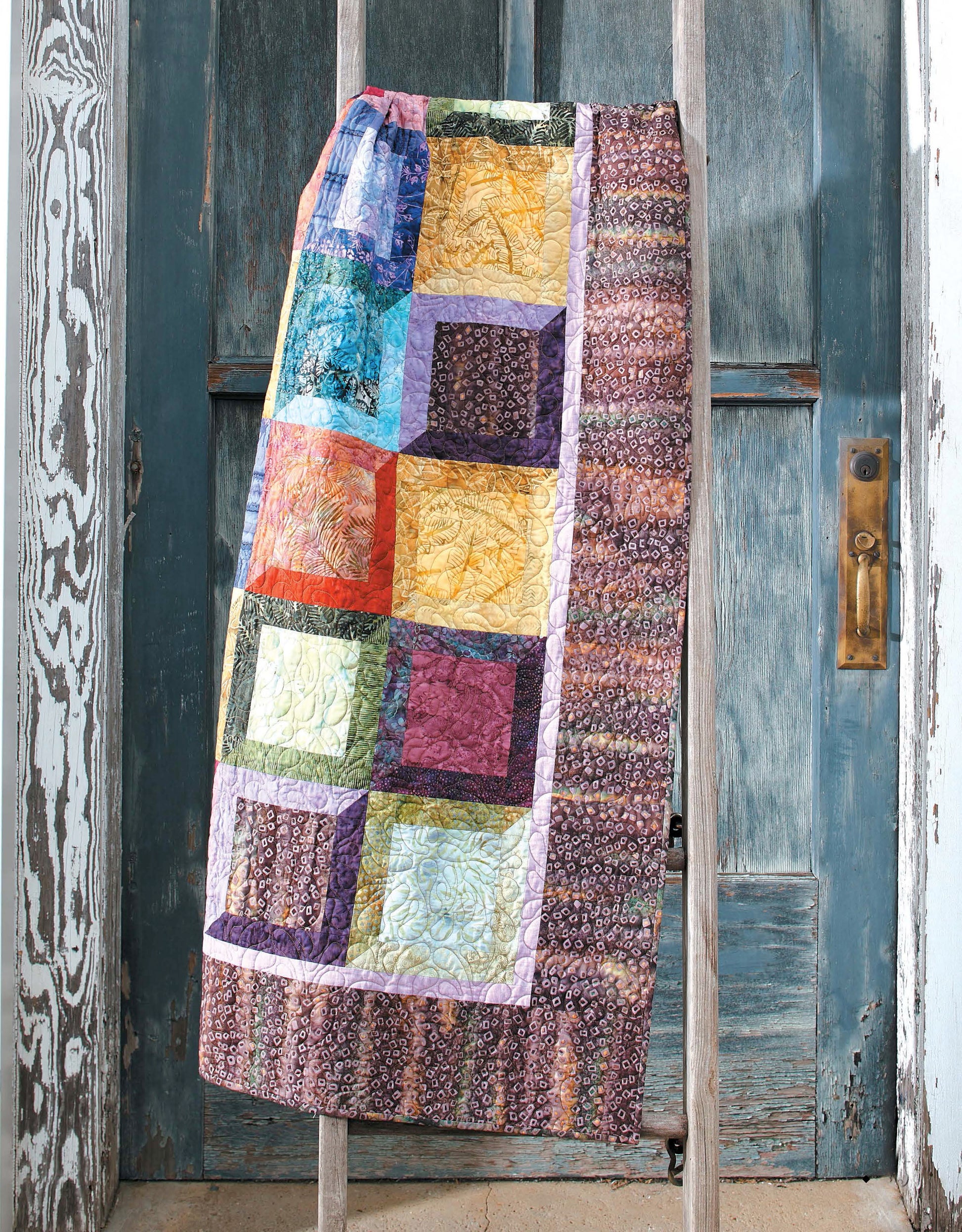 A vibrant quilt from Leisure Arts’ "Fresh, Fast & Fun Quilts," made with fat quarter techniques and patterned squares, is draped over a wooden ladder leaning against a weathered blue door with peeling paint and a brass handle.