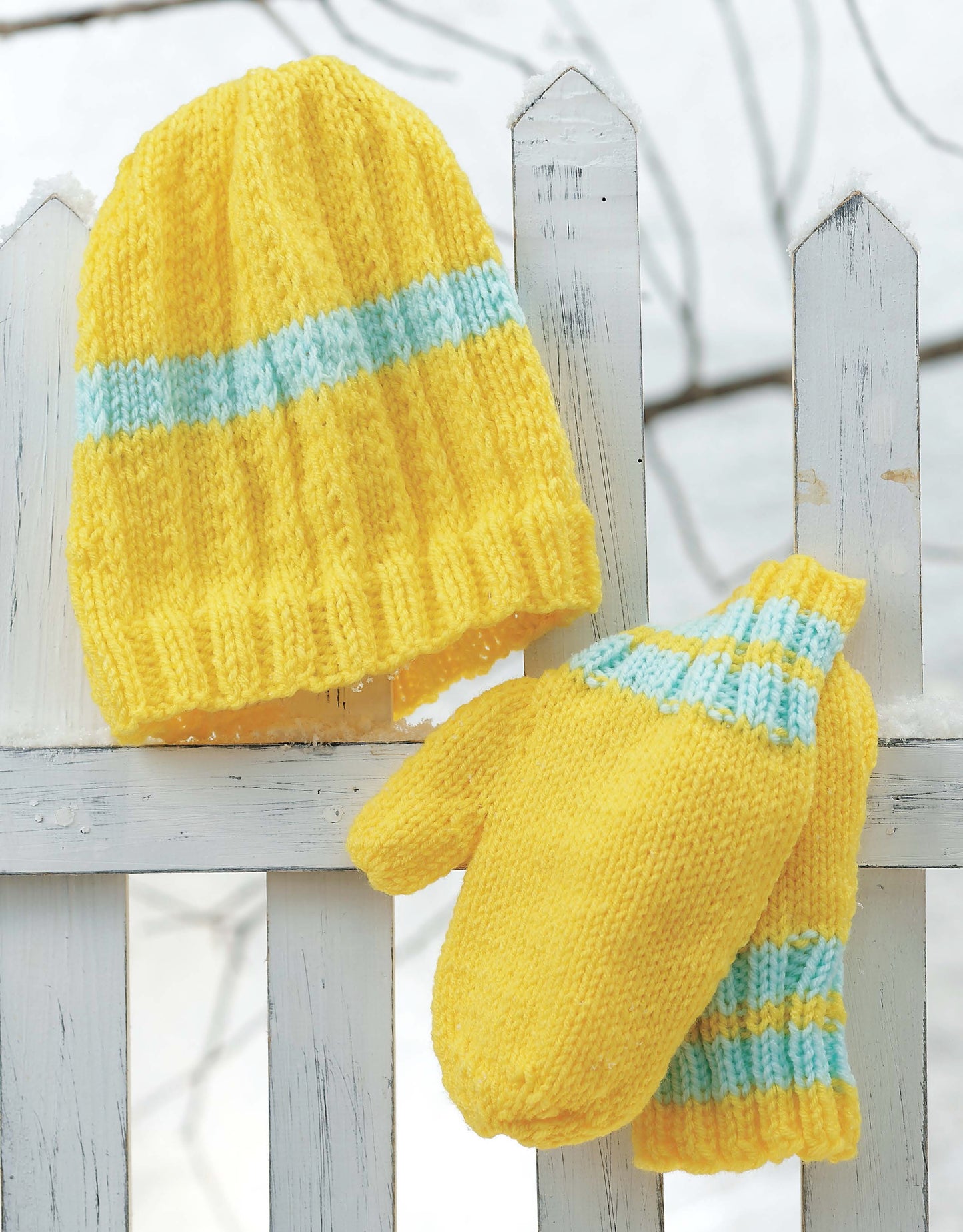 The Snow Day Sets to Knit by Leisure Arts features a yellow knitted hat and matching mittens with blue and white stripes, displayed on a white picket fence against a snowy backdrop.