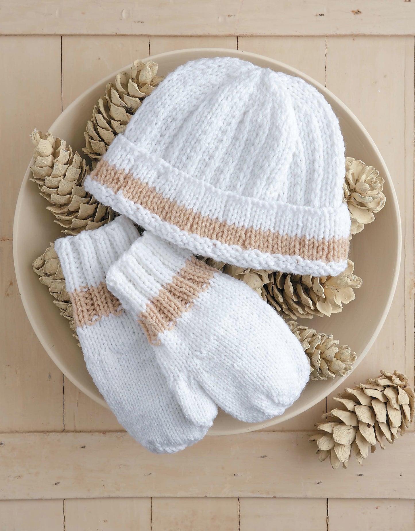 The Snow Day Sets to Knit by Leisure Arts features a white hat and matching mittens with beige stripes, crafted from worsted weight yarn, displayed alongside white pinecones on a light wooden surface.