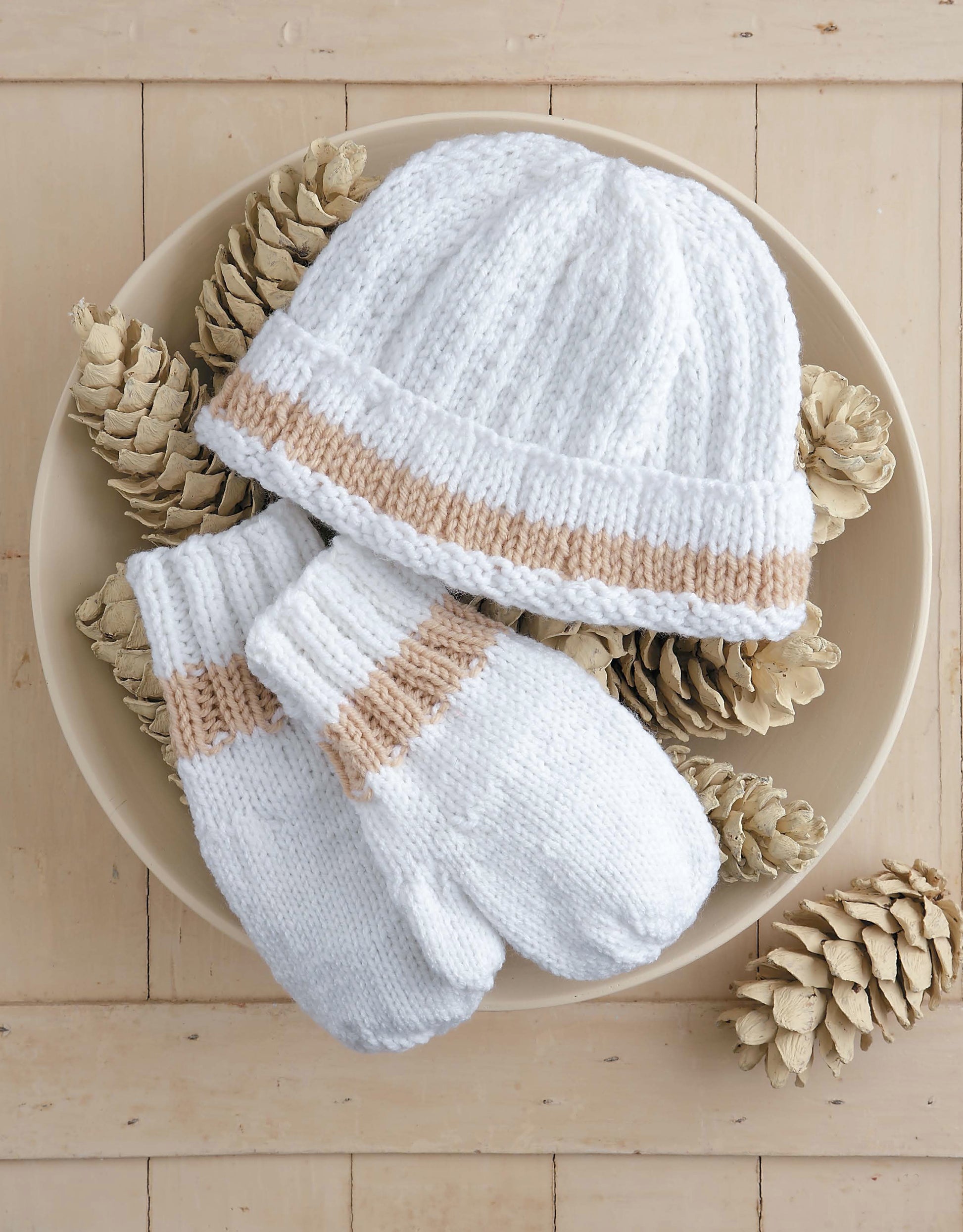 The Snow Day Sets to Knit by Leisure Arts features a white hat and matching mittens with beige stripes, crafted from worsted weight yarn, displayed alongside white pinecones on a light wooden surface.