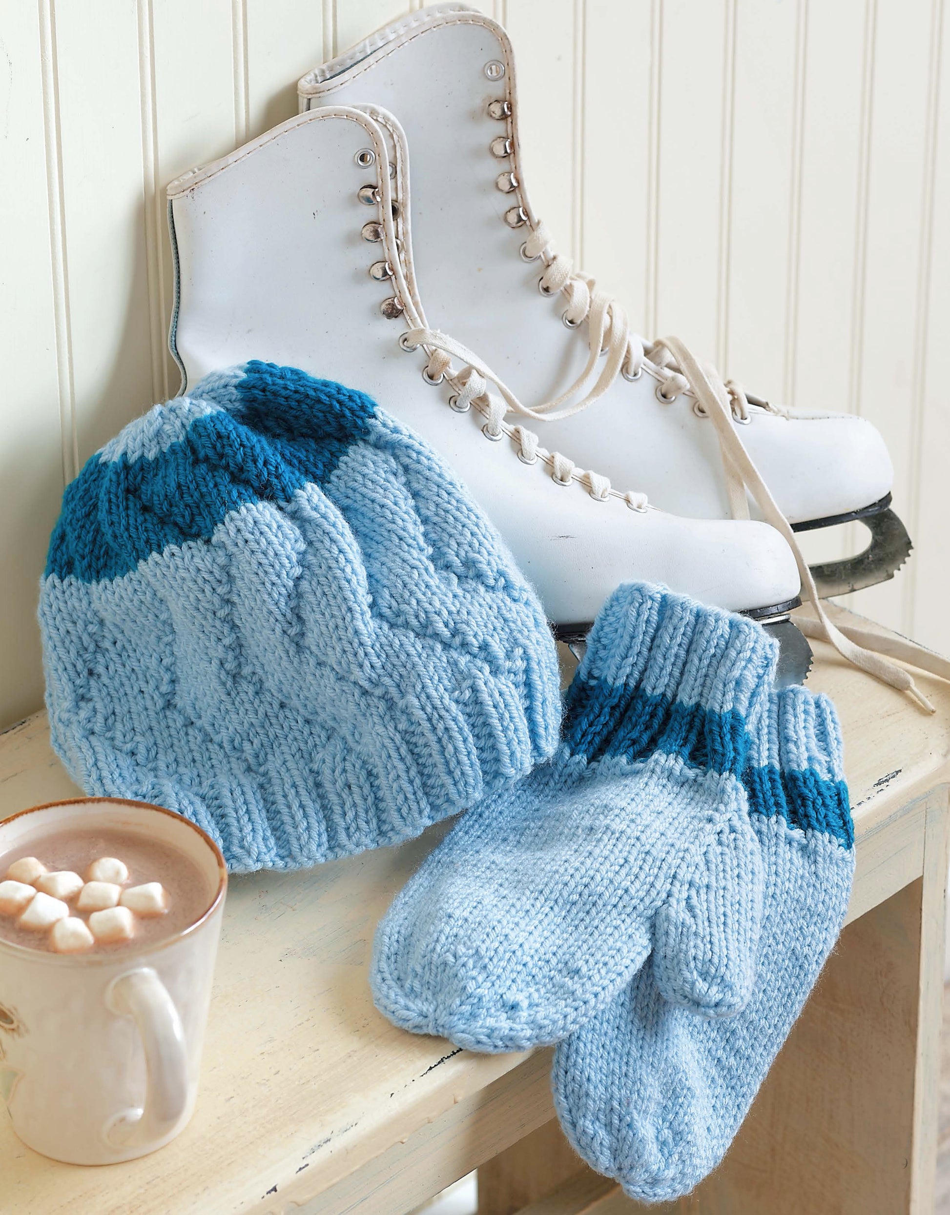 The Snow Day Sets to Knit by Leisure Arts features a light blue worsted-weight hat with matching mittens accented in dark blue, white ice skates, and a hot chocolate mug with marshmallows on a wooden bench.