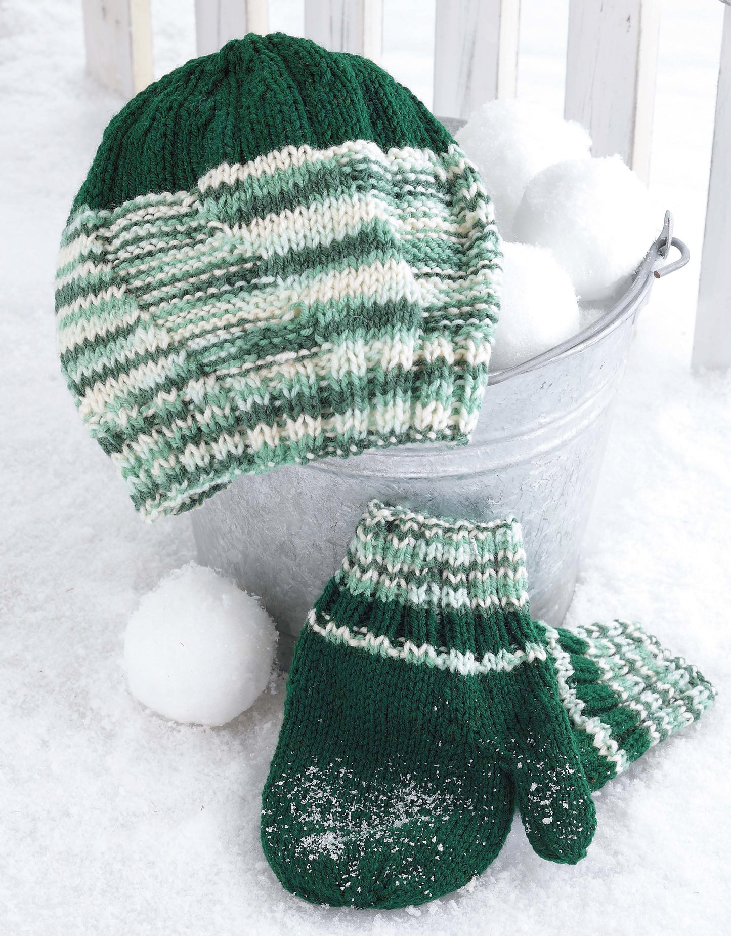 The Snow Day Sets to Knit by Leisure Arts features a green and white striped knit hat and matching mittens in worsted weight yarn, resting on snow beside a metal bucket of snowballs, with the mittens lightly dusted with snow.