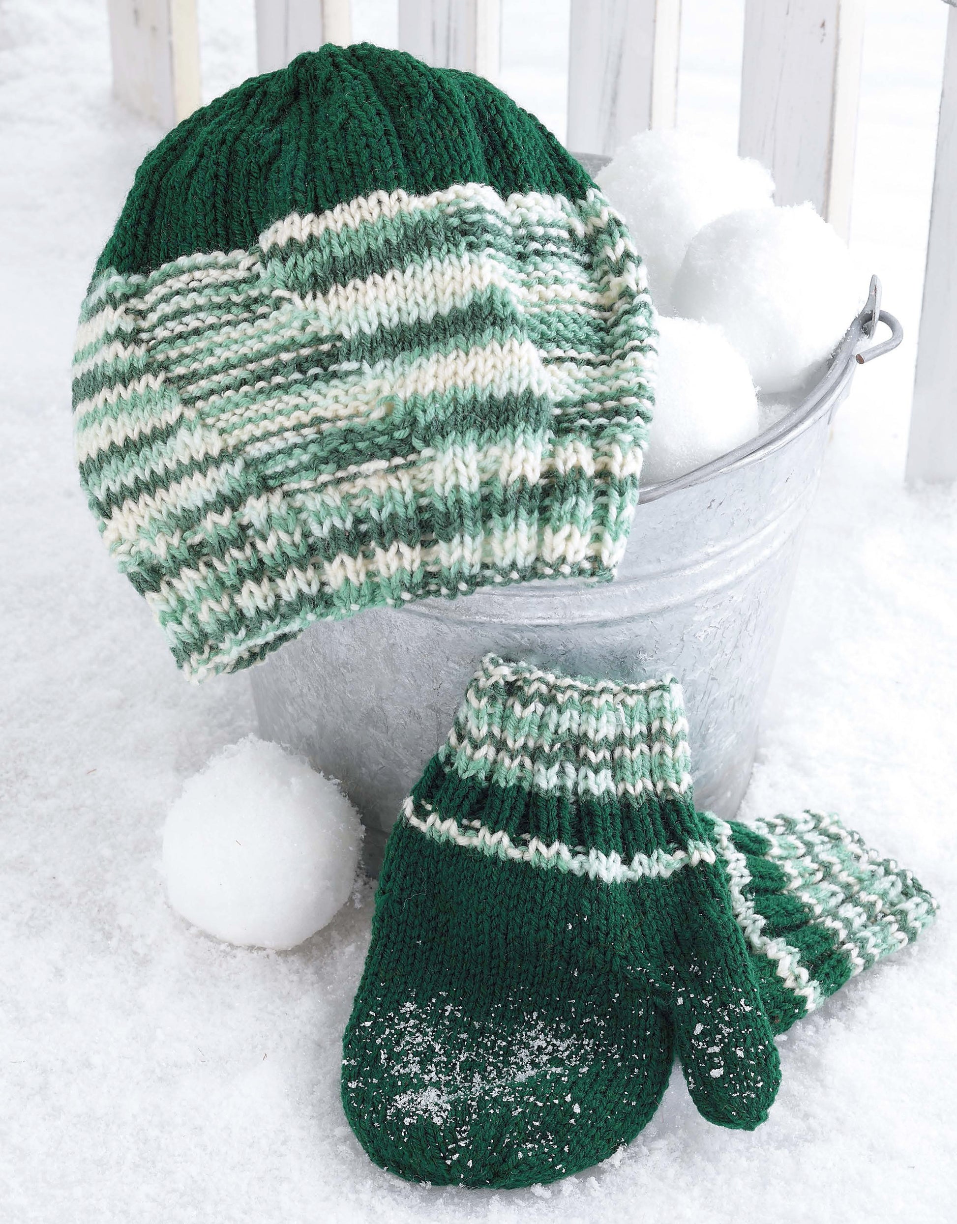 The Snow Day Sets to Knit by Leisure Arts features a green and white striped knit hat and matching mittens in worsted weight yarn, resting on snow beside a metal bucket of snowballs, with the mittens lightly dusted with snow.