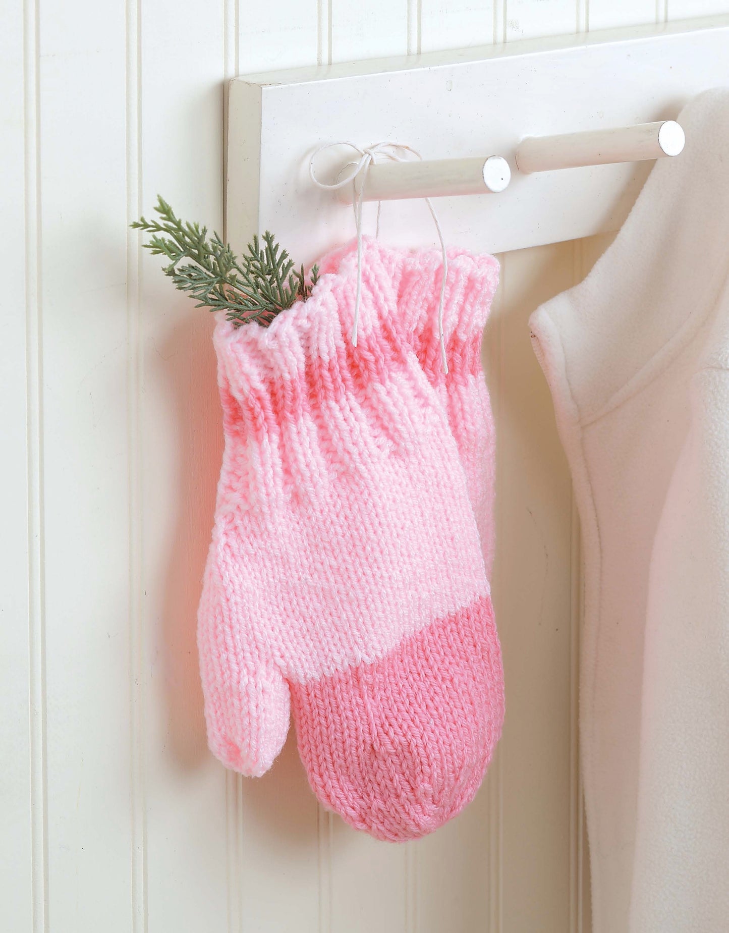 A pair of pink mittens, made from worsted yarn using Leisure Arts' Snow Day Sets to Knit, hangs on a white hook with a green sprig. A white garment hangs nearby against a white paneled wall.