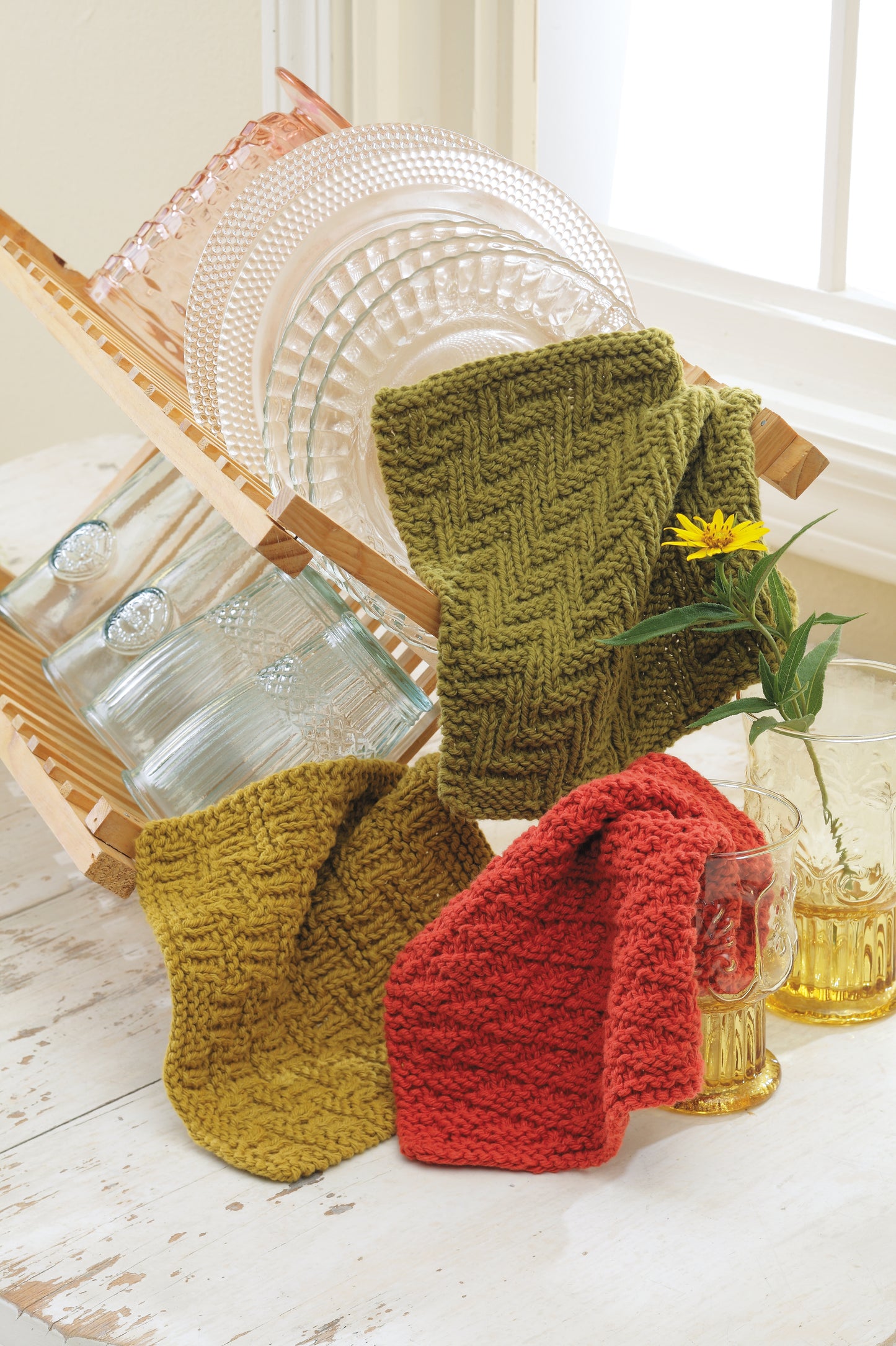 Three textured knitted dishcloths from Leisure Arts’ Easy Textured Knits Digital Download in olive green, chartreuse, and red-orange are displayed on a wooden rack with glass plates, amber glasses, and a yellow flower on a white table.