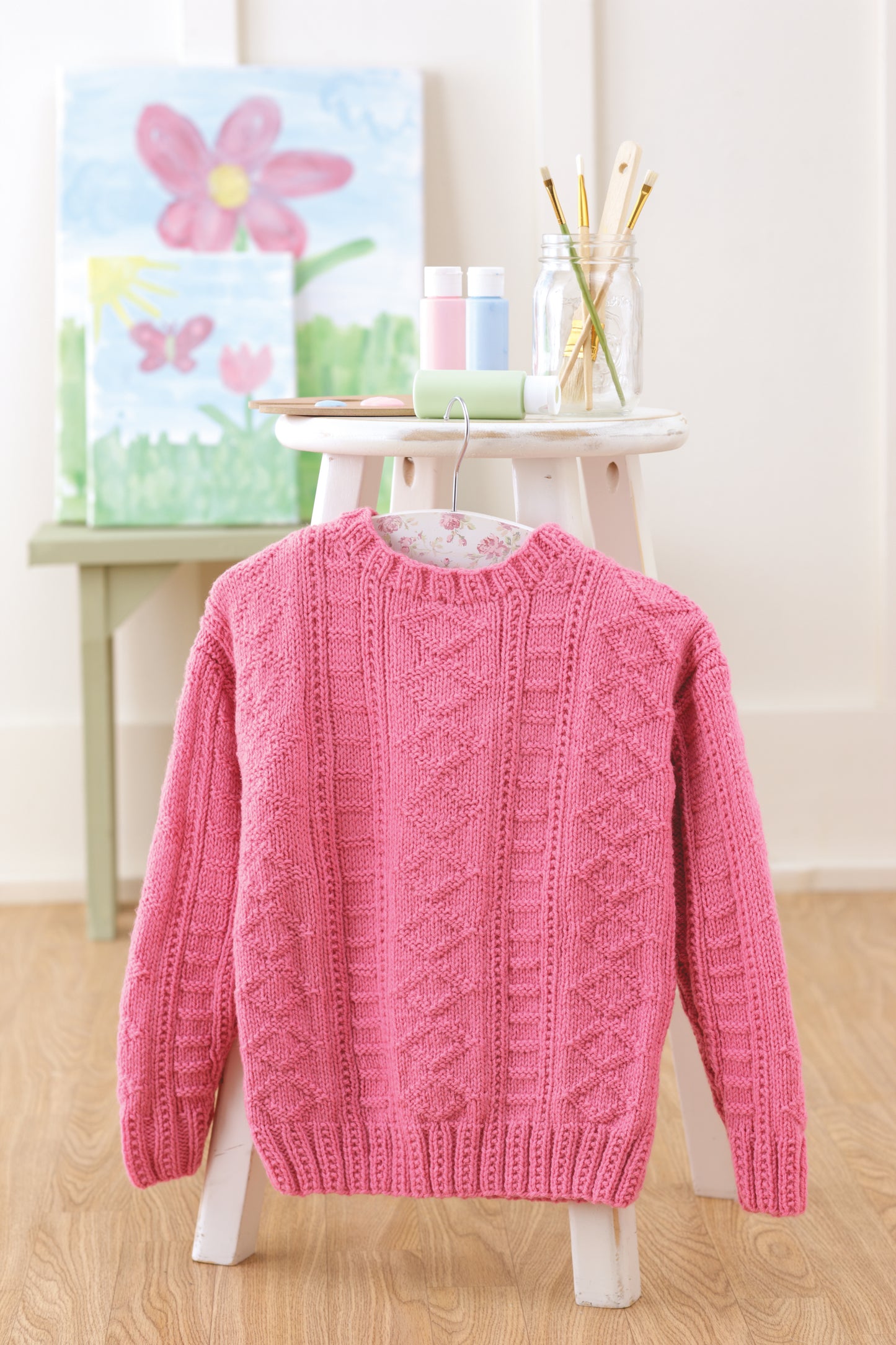 The Easy Textured Knits Digital Download by Leisure Arts is displayed on a pink sweater draped over a white wooden stool, surrounded by art supplies and pastel decor, in a bright, cozy crafting space.