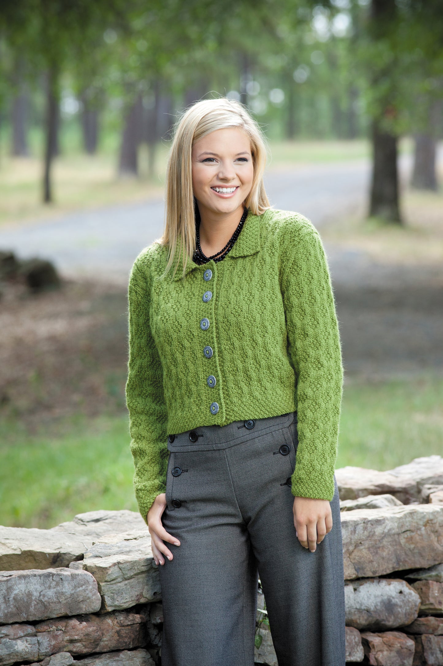 A smiling woman with straight blonde hair models a green textured button-up sweater from Leisure Arts' Easy Textured Knits Digital Download, paired with gray high-waisted pants, standing outdoors by a stone wall and trees.