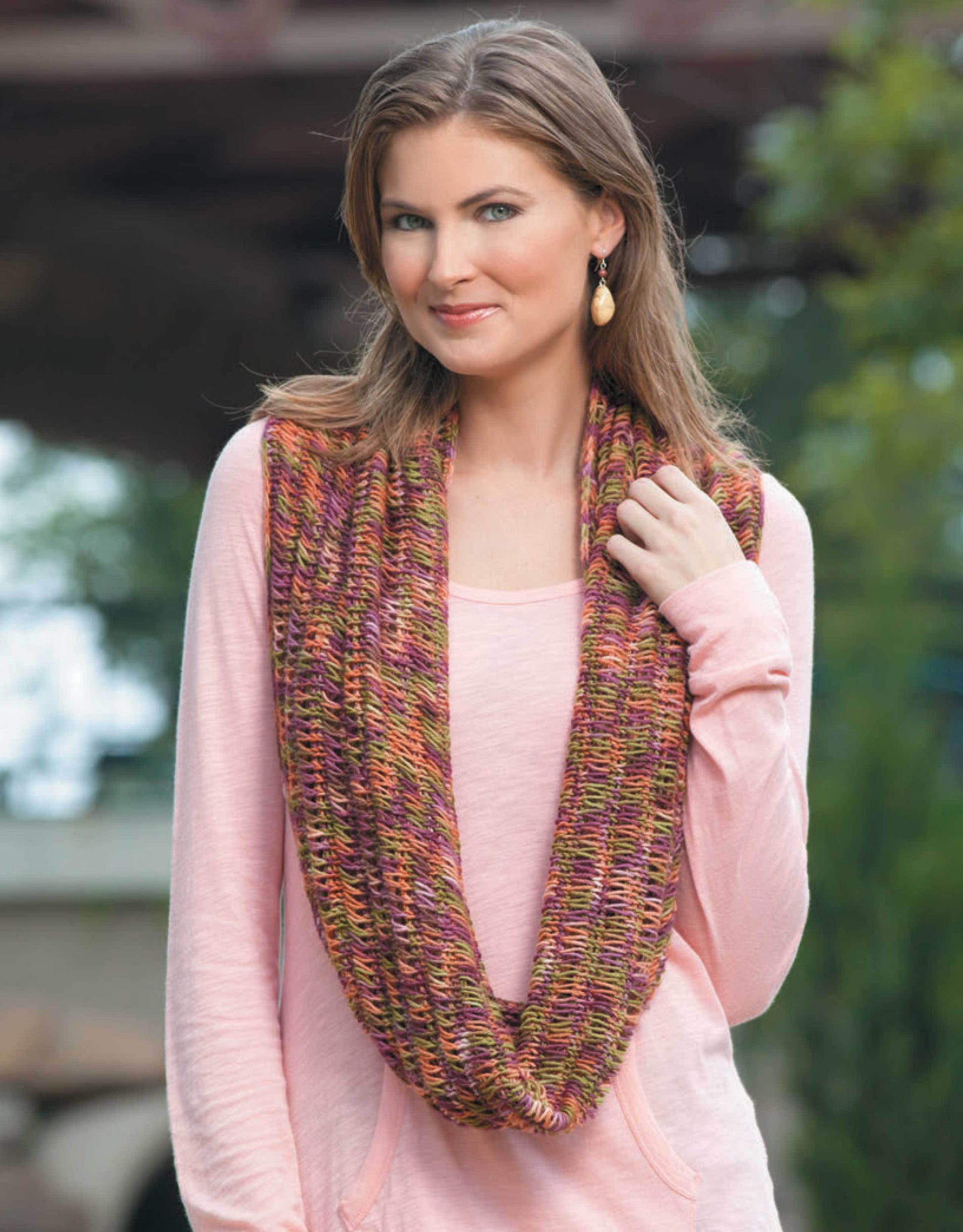 A woman with light brown hair wears a pink long-sleeve top and a colorful knit scarf—ideal for beginner projects featured in Leisure Arts' "Ultimate Beginner's Guide to Tunisian Crochet"—as she smiles gently outdoors.