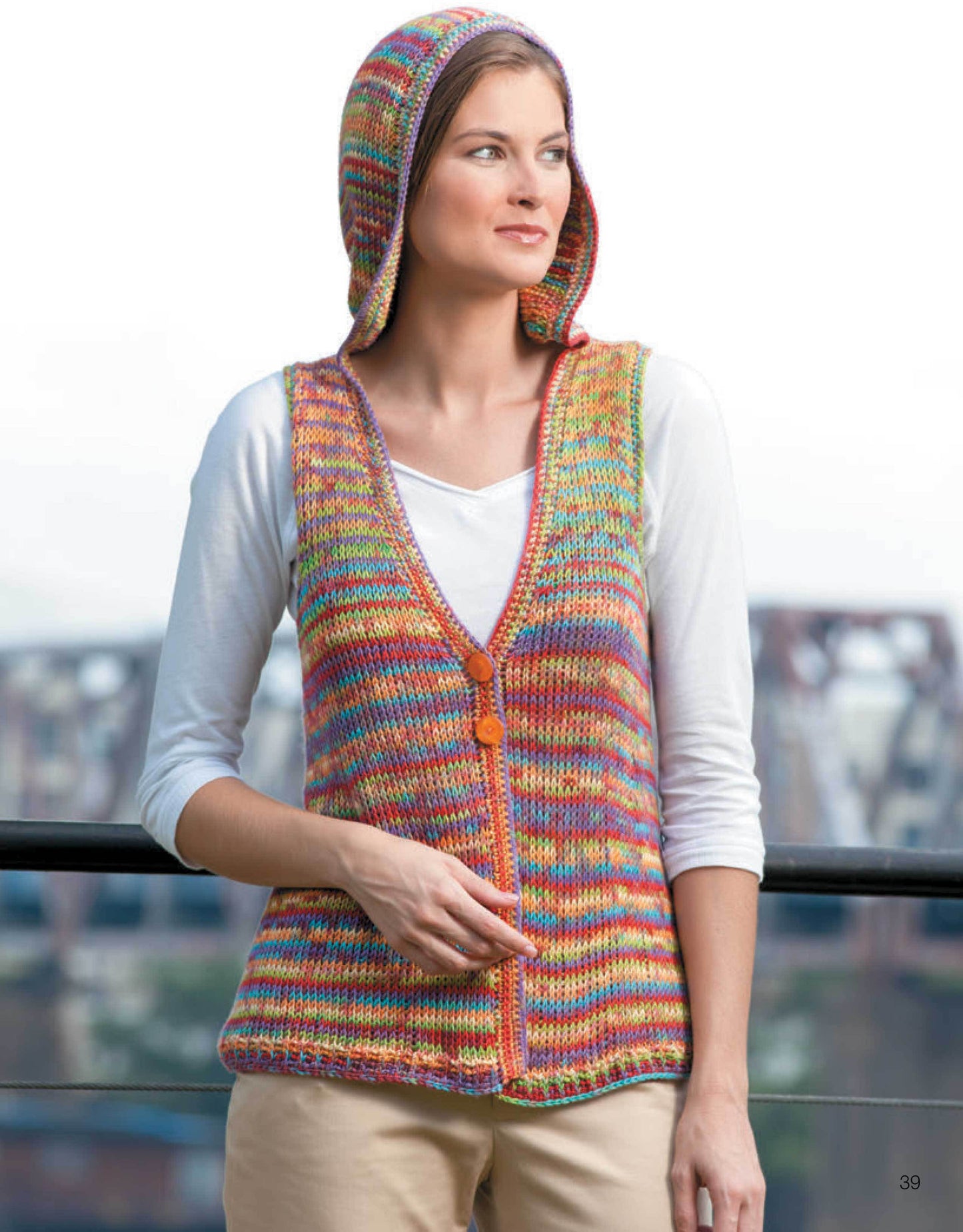A woman stands outdoors in a colorful, sleeveless, hooded knit vest made with medium weight yarn over a white shirt. She rests a hand on a railing; the scene promotes Leisure Arts’ "Ultimate Beginner's Guide to Tunisian Crochet.