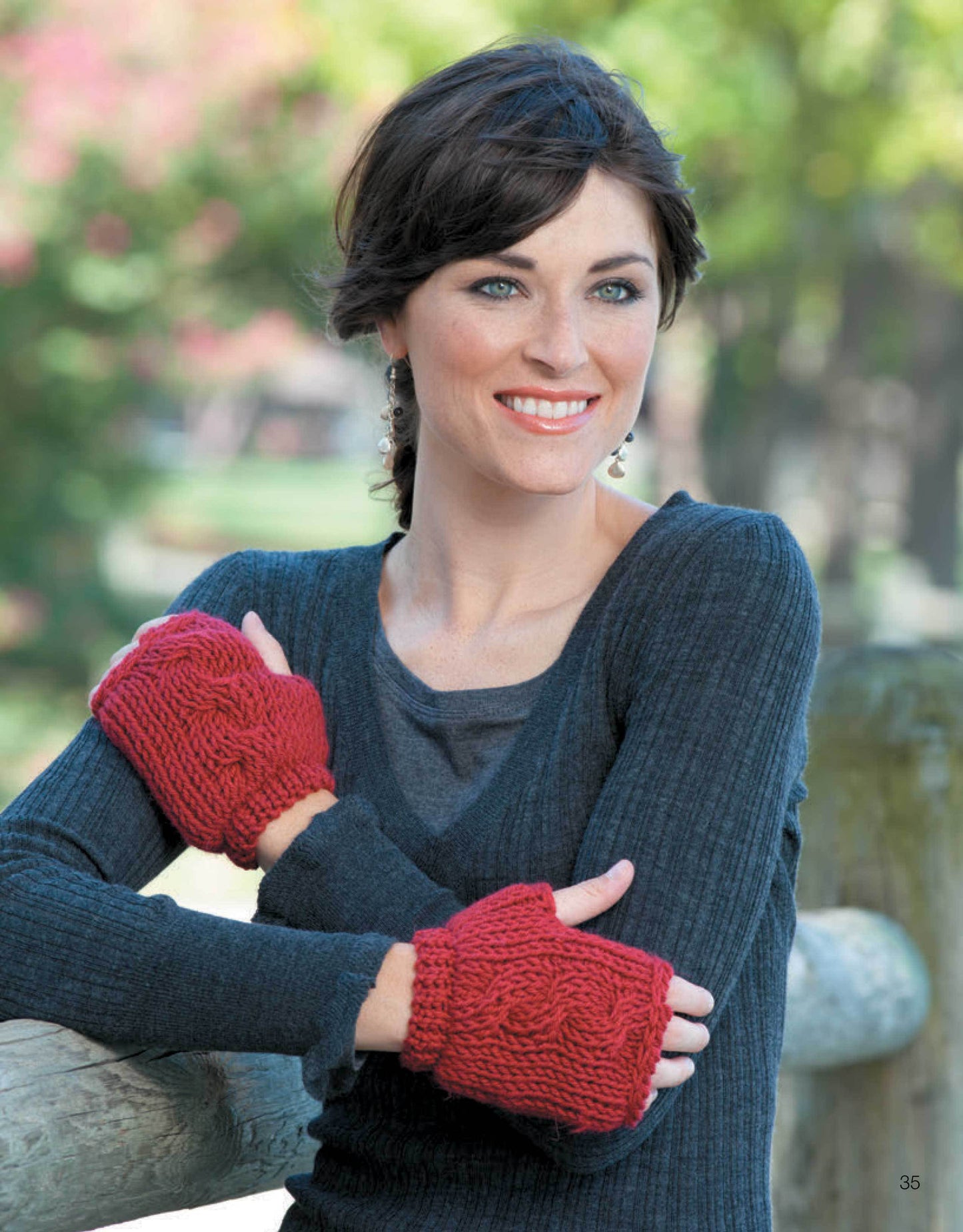 A woman with short dark hair smiles outdoors in a dark long-sleeve top, red fingerless gloves, and a chunky scarf—great for beginner projects inspired by Leisure Arts’ Ultimate Beginner's Guide to Tunisian Crochet. She leans on a wooden fence.