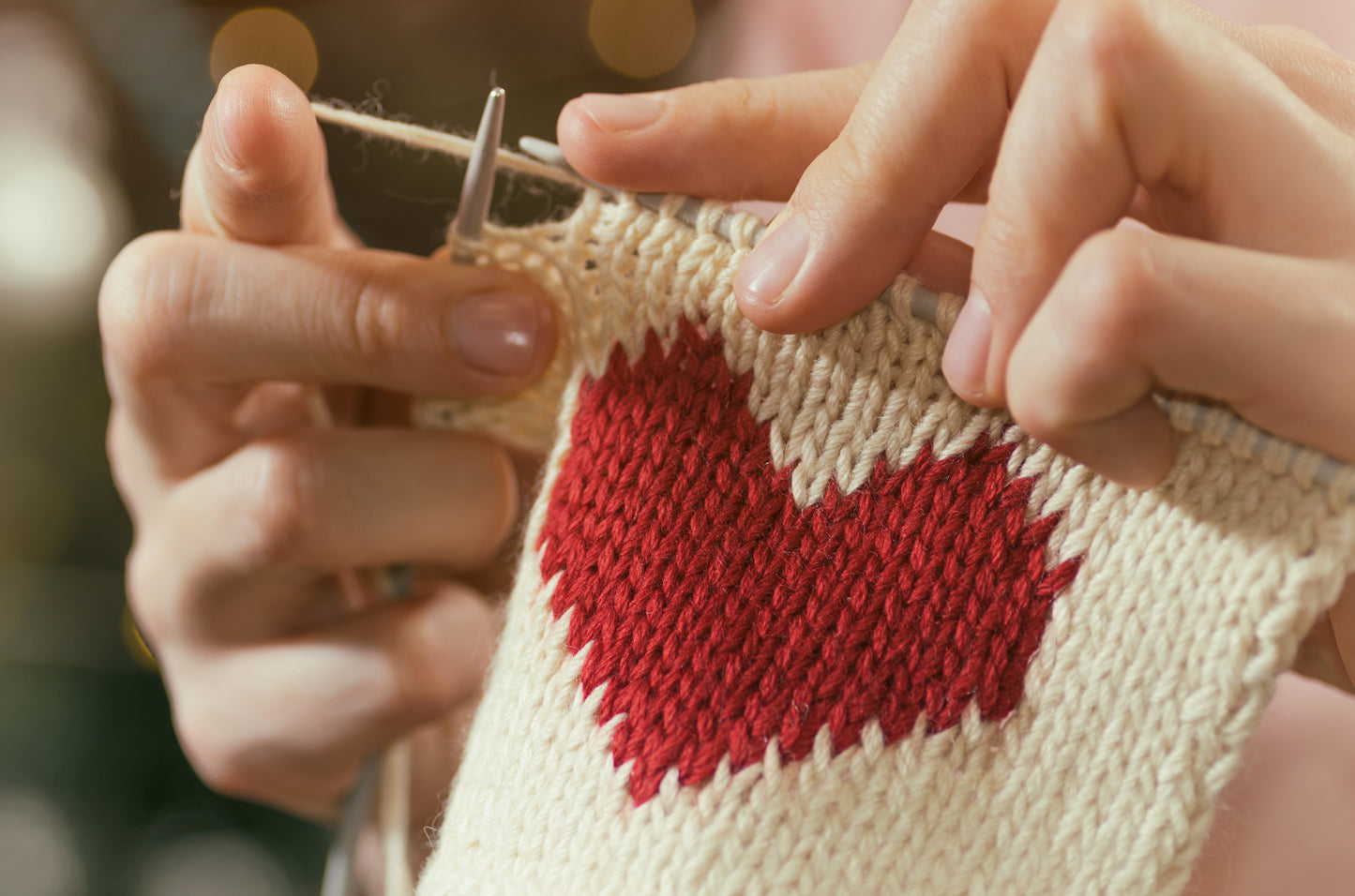 Close-up of hands knitting a cream fabric with a red heart using needles, ideal for beginners. Features patterns inspired by Leisure Arts and techniques from the Knitting Pocket Guide. Blurred background lights add a cozy touch.