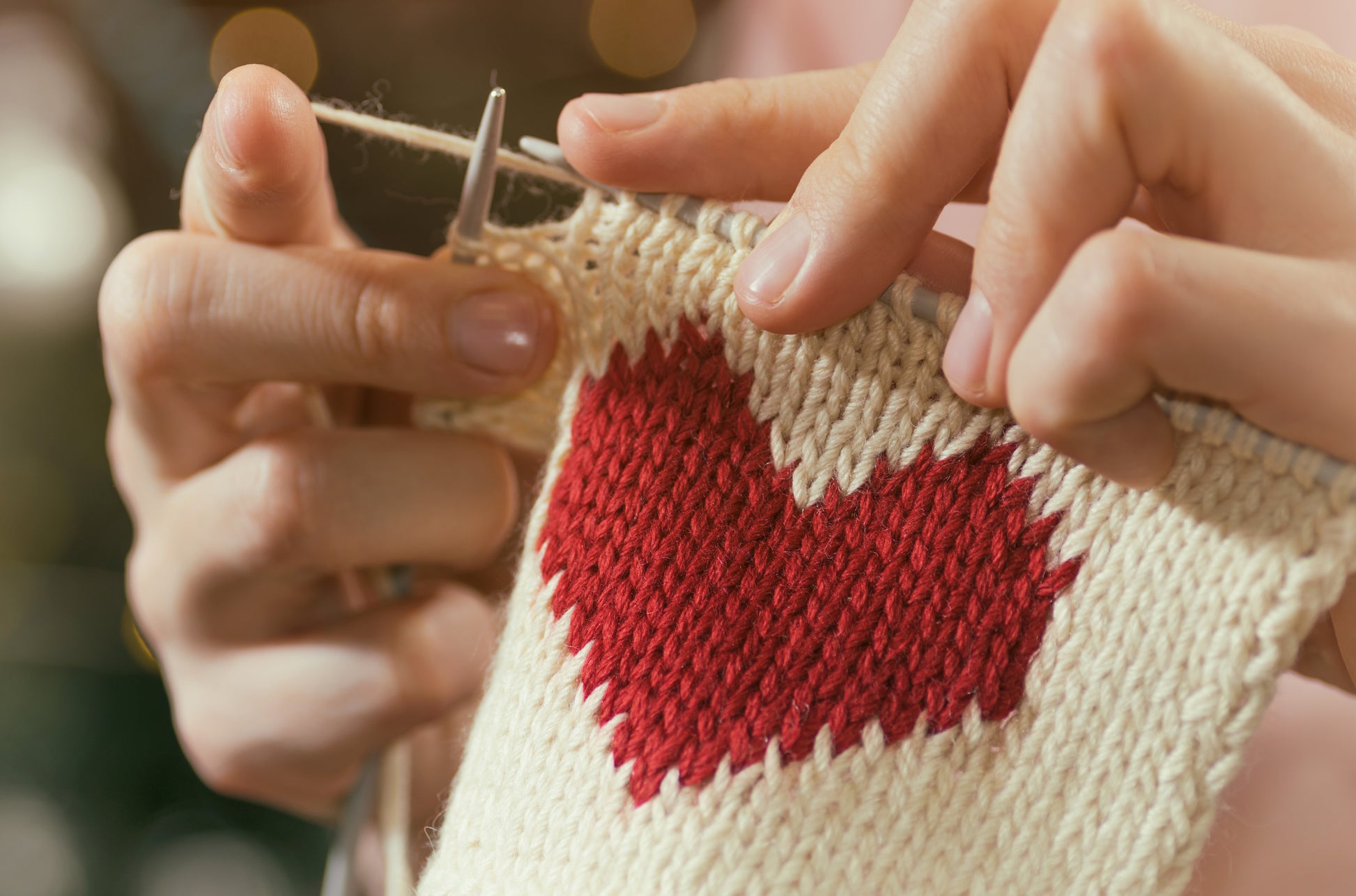 Close-up of hands knitting a cream fabric with a red heart using needles, ideal for beginners. Features patterns inspired by Leisure Arts and techniques from the Knitting Pocket Guide. Blurred background lights add a cozy touch.