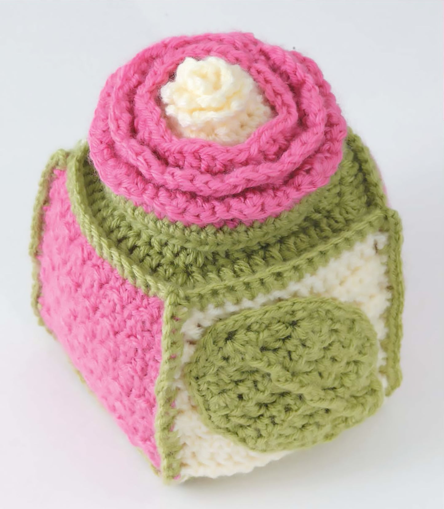 The Leisure Arts "Crochet Textures for Home & Baby" features a soft cube made with worsted weight yarn, pink sides, green accents, and a spiral top resembling a cake, displayed against a plain white background.
