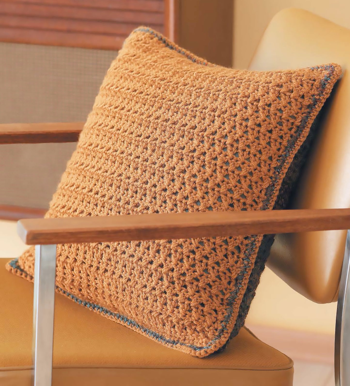 A square, orange pillow with a sampler-inspired crochet texture and blue trim from Leisure Arts' "Crochet Textures for Home & Baby" sits on a light tan, mid-century modern chair with wooden armrests.
