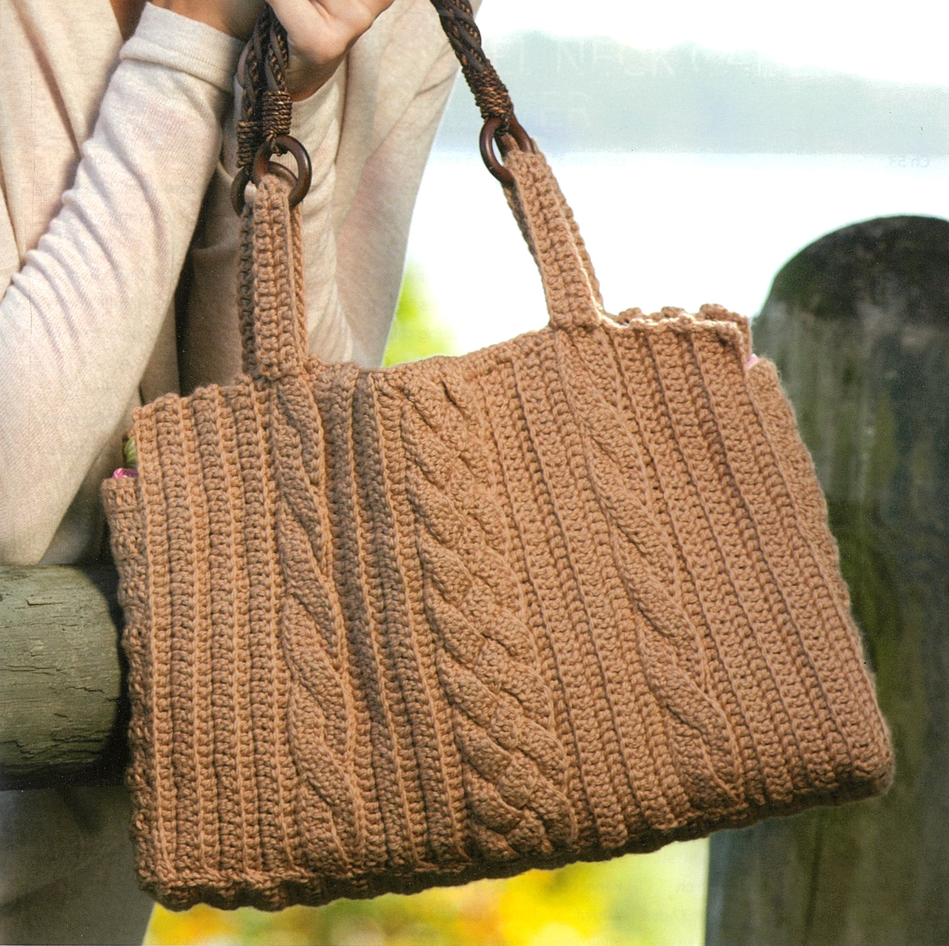 A person holds a brown handbag made with Add-On Crochet Cables by Leisure Arts, featuring two handles and textured crochet cables. The accessory is shown outdoors near a wooden post, with part of a beige sleeve also visible.