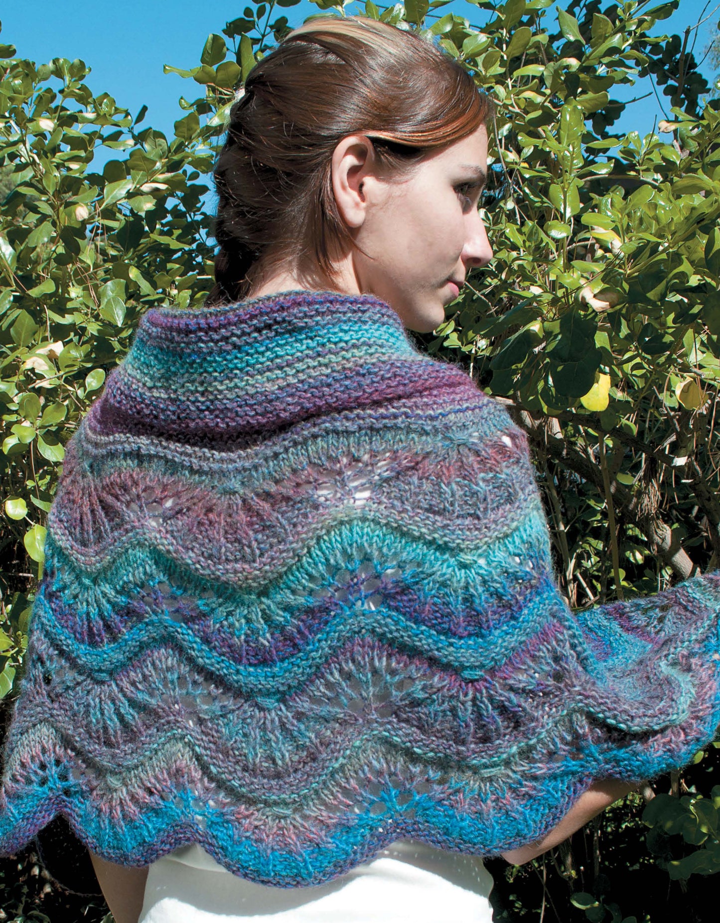 A woman with brown hair pulled back stands outdoors before green bushes, modeling a vibrant shawl knit in waves of blue, purple, and teal—an eye-catching piece from Leisure Arts’ "Shawls, Wraps, & Ponchos to Knit.