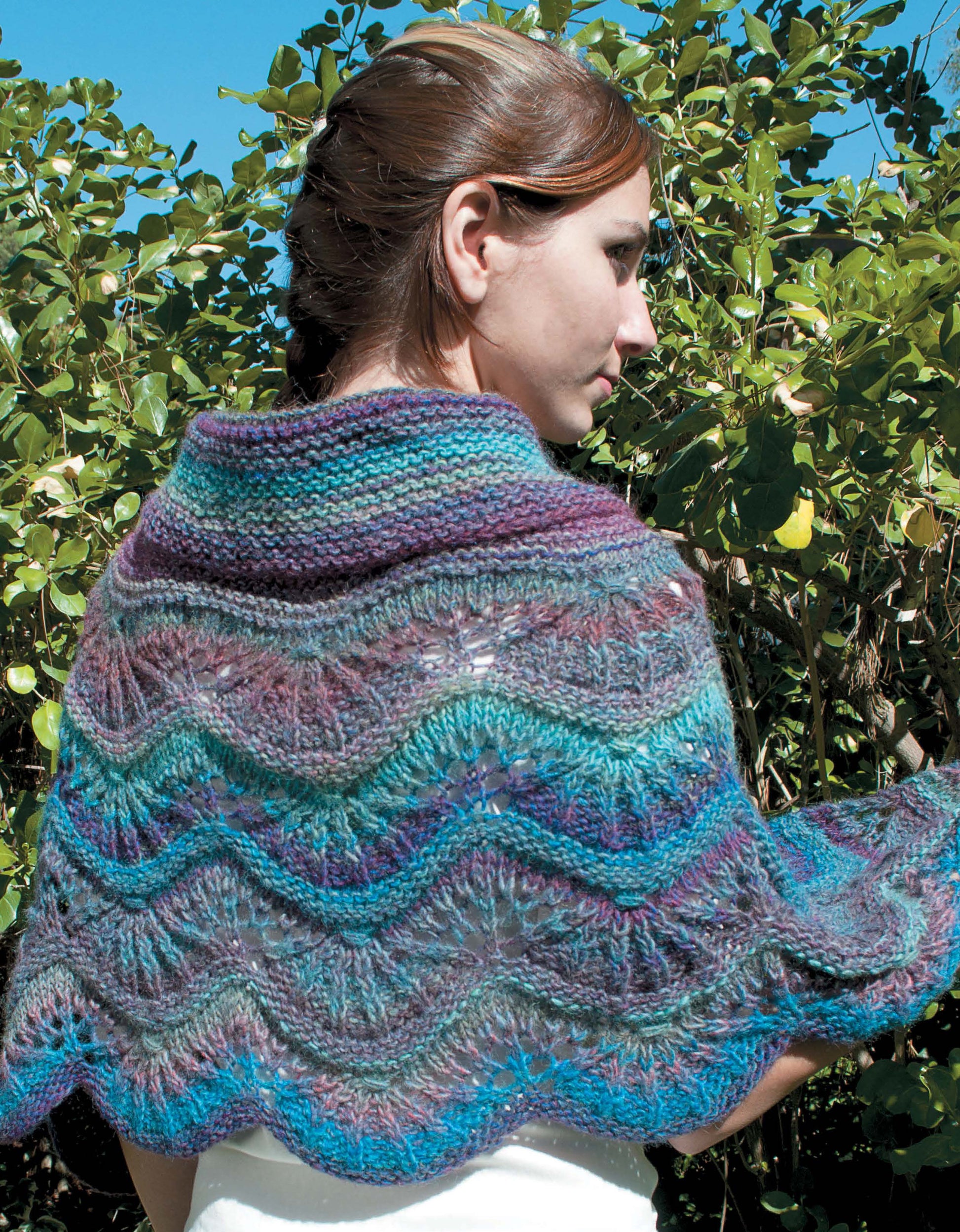 A woman with brown hair pulled back stands outdoors before green bushes, modeling a vibrant shawl knit in waves of blue, purple, and teal—an eye-catching piece from Leisure Arts’ "Shawls, Wraps, & Ponchos to Knit.