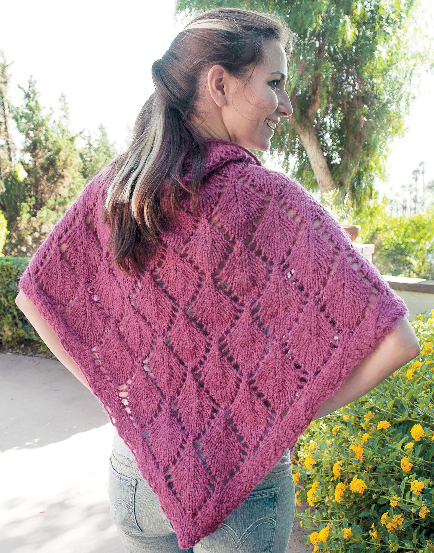 A woman outdoors wears a purple, lacy knit shawl from "Shawls, Wraps, & Ponchos to Knit" by Leisure Arts. Draped over her shoulders, she stands among trees and flowers, smiling with her back to the camera.