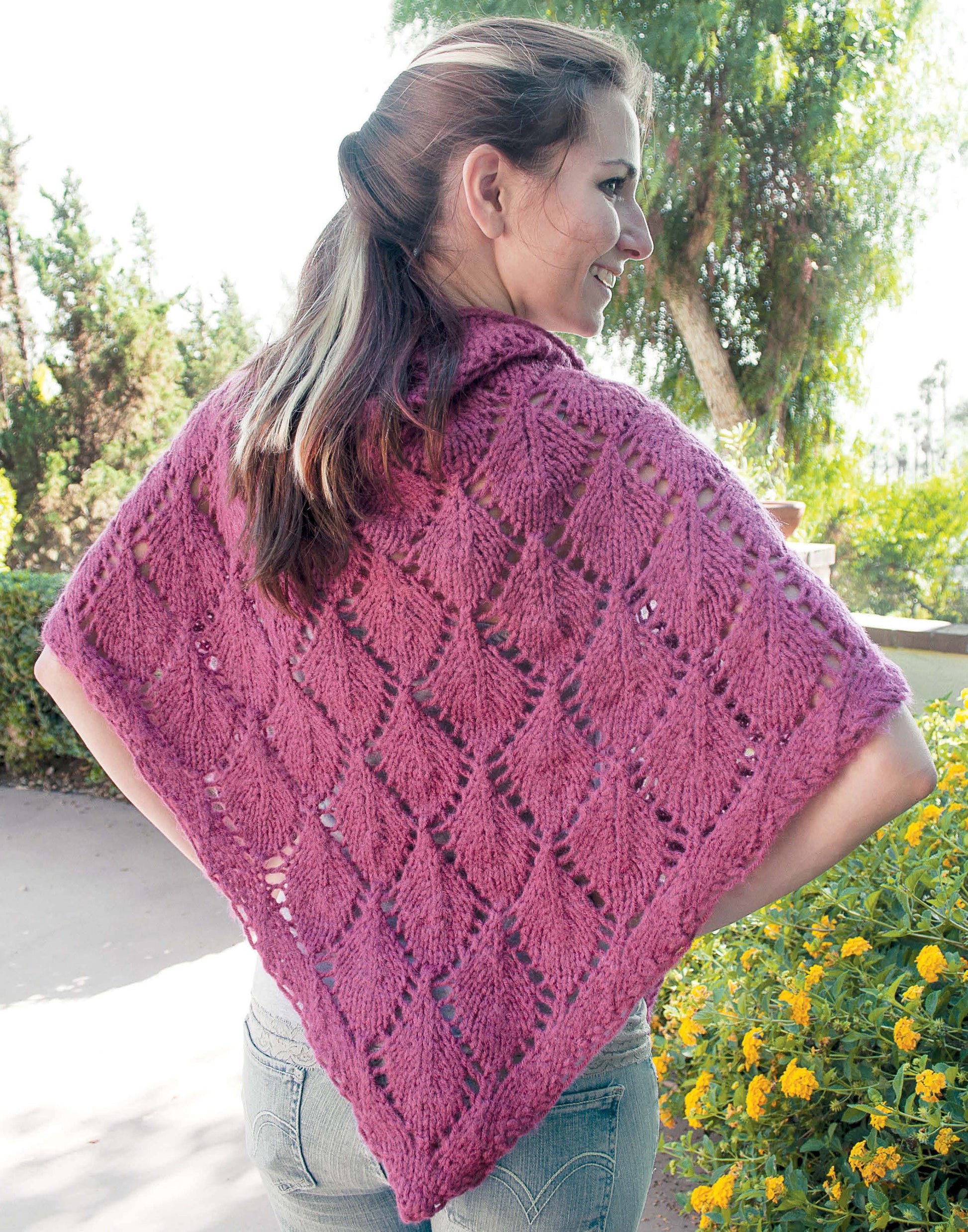 A woman outdoors wears a purple, lacy knit shawl from "Shawls, Wraps, & Ponchos to Knit" by Leisure Arts. Draped over her shoulders, she stands among trees and flowers, smiling with her back to the camera.