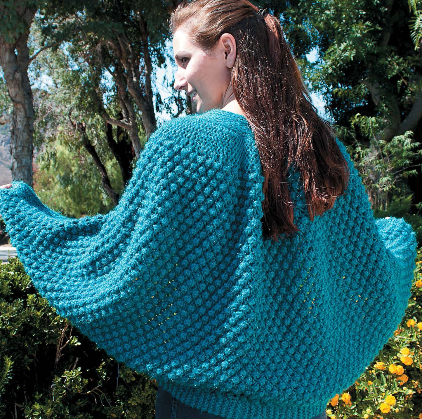 A woman outdoors wears a teal hand-knitted shrug, made with patterns from "Shawls, Wraps, & Ponchos to Knit" by Leisure Arts. She stands among sunny green bushes and trees, her long brown hair catching the light.