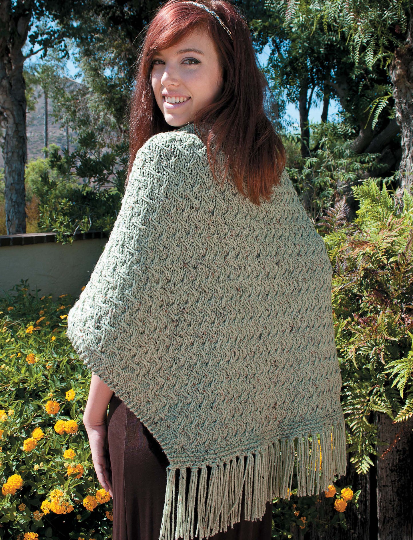 A woman with reddish-brown hair stands outdoors among yellow flowers, smiling while wearing a light green textured shawl made from Leisure Arts' "Shawls, Wraps, & Ponchos to Knit.