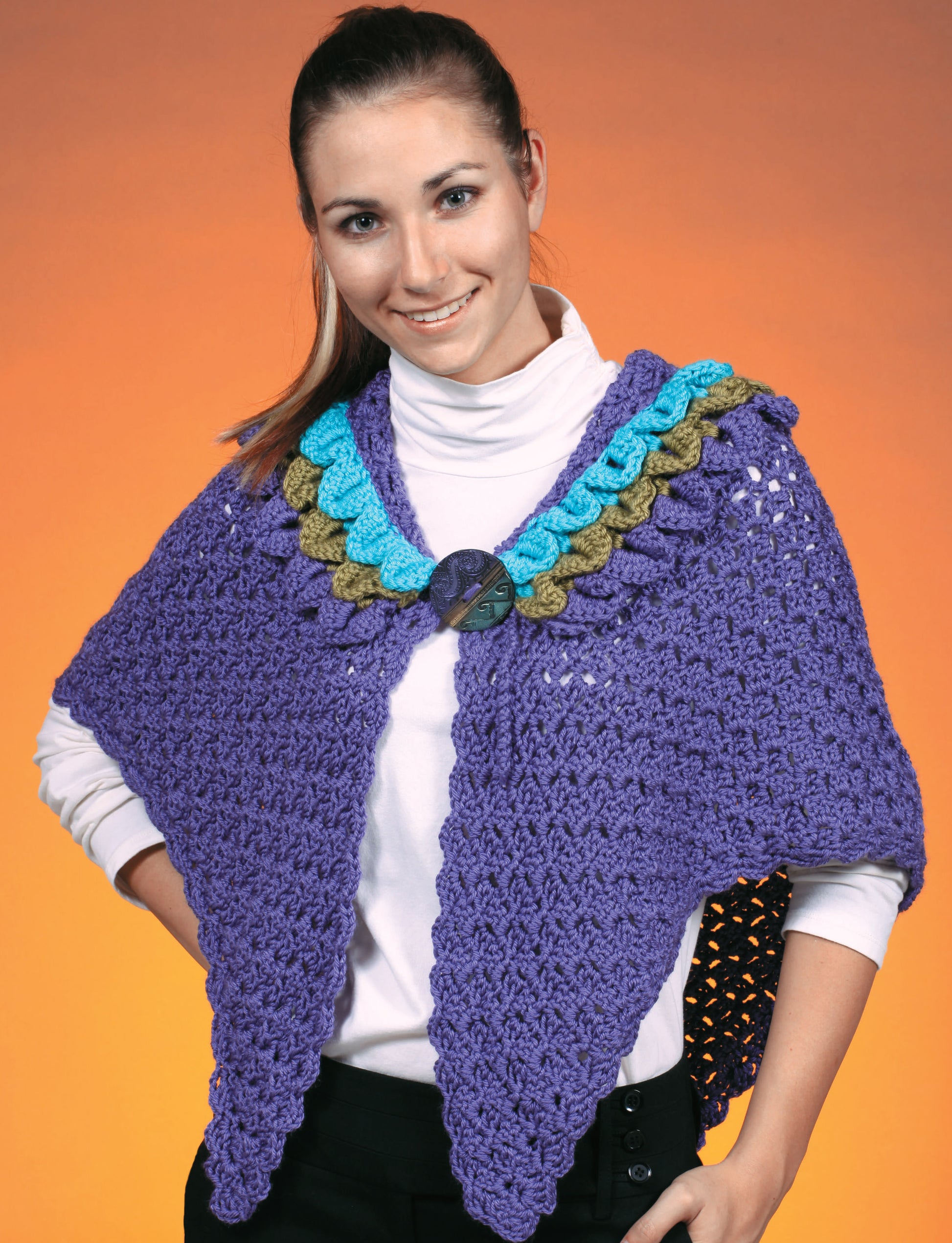 Model wears a purple crocheted shawl with blue, teal, and olive green scalloped border from "Make in a Weekend Shawls" by Leisure Arts, styled over a white turtleneck and black pants against an orange background.