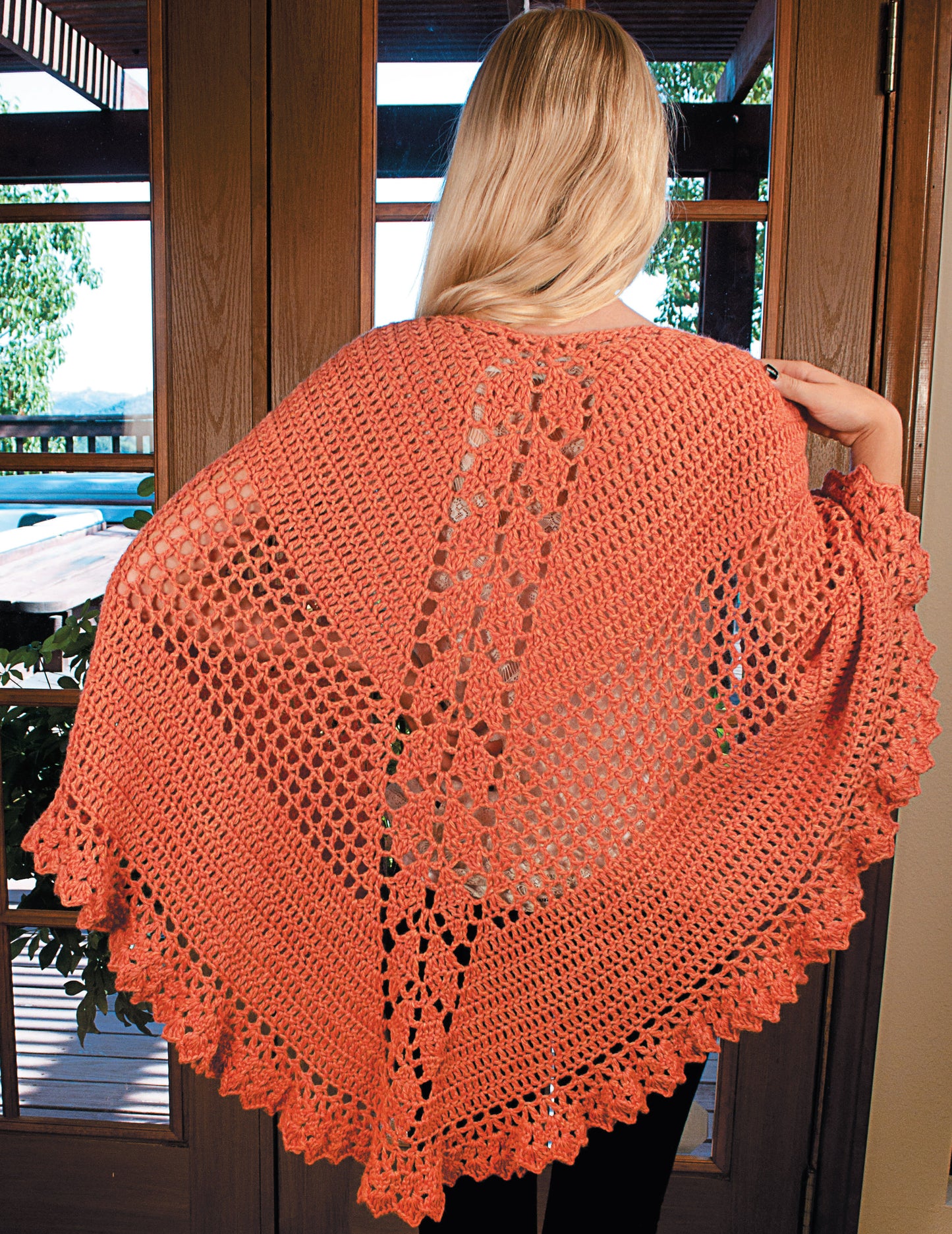 A woman with long blonde hair models a coral floral crochet shawl from Make in a Weekend Shawls by Leisure Arts, her back to the camera indoors near wooden doors—a stylish pattern suitable for all ages.