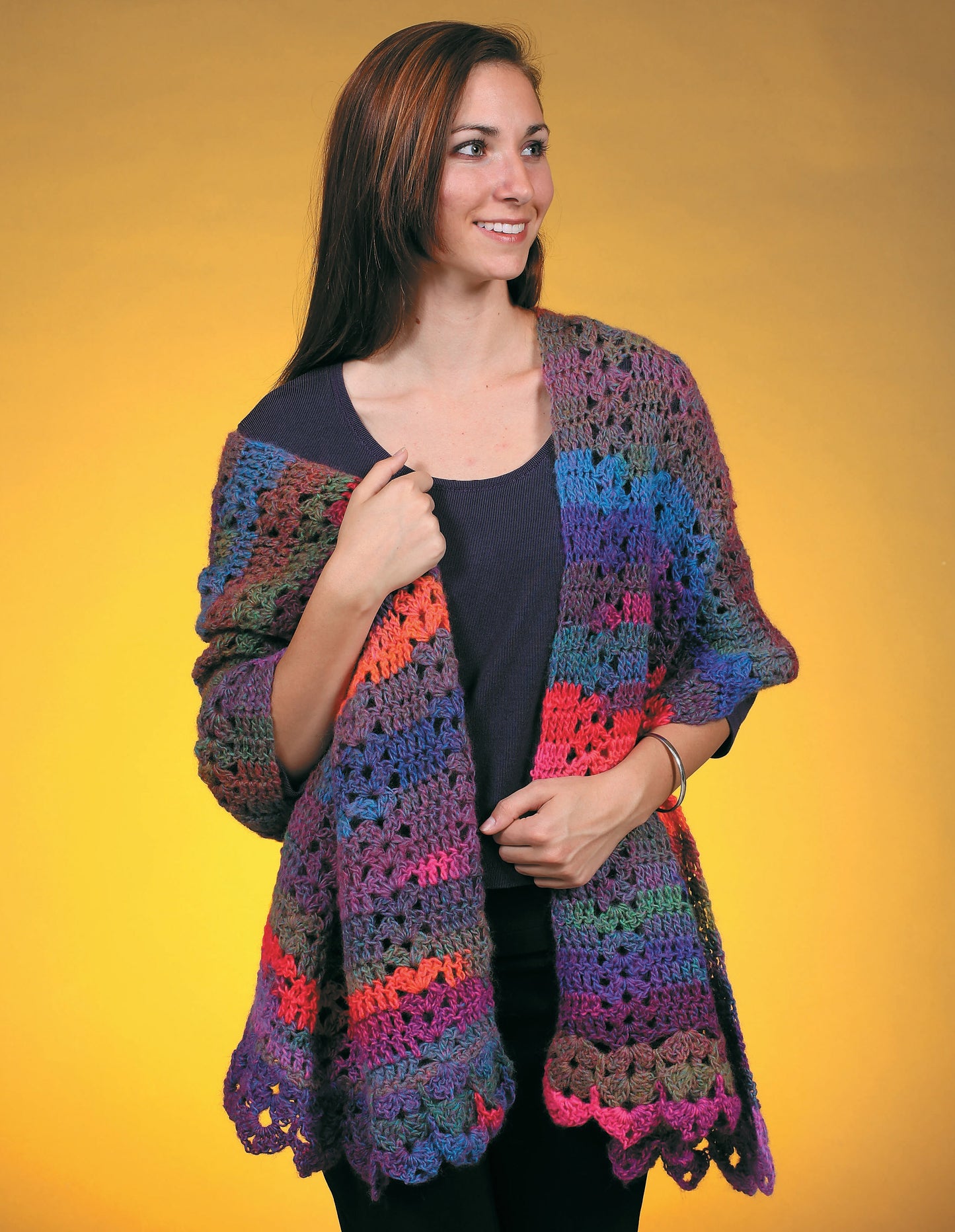 A woman with long brown hair smiles while wearing a colorful crocheted shawl from Leisure Arts' "Make in a Weekend Shawls," over a dark shirt, standing against a yellow background. Perfect for those seeking crochet shawl patterns.