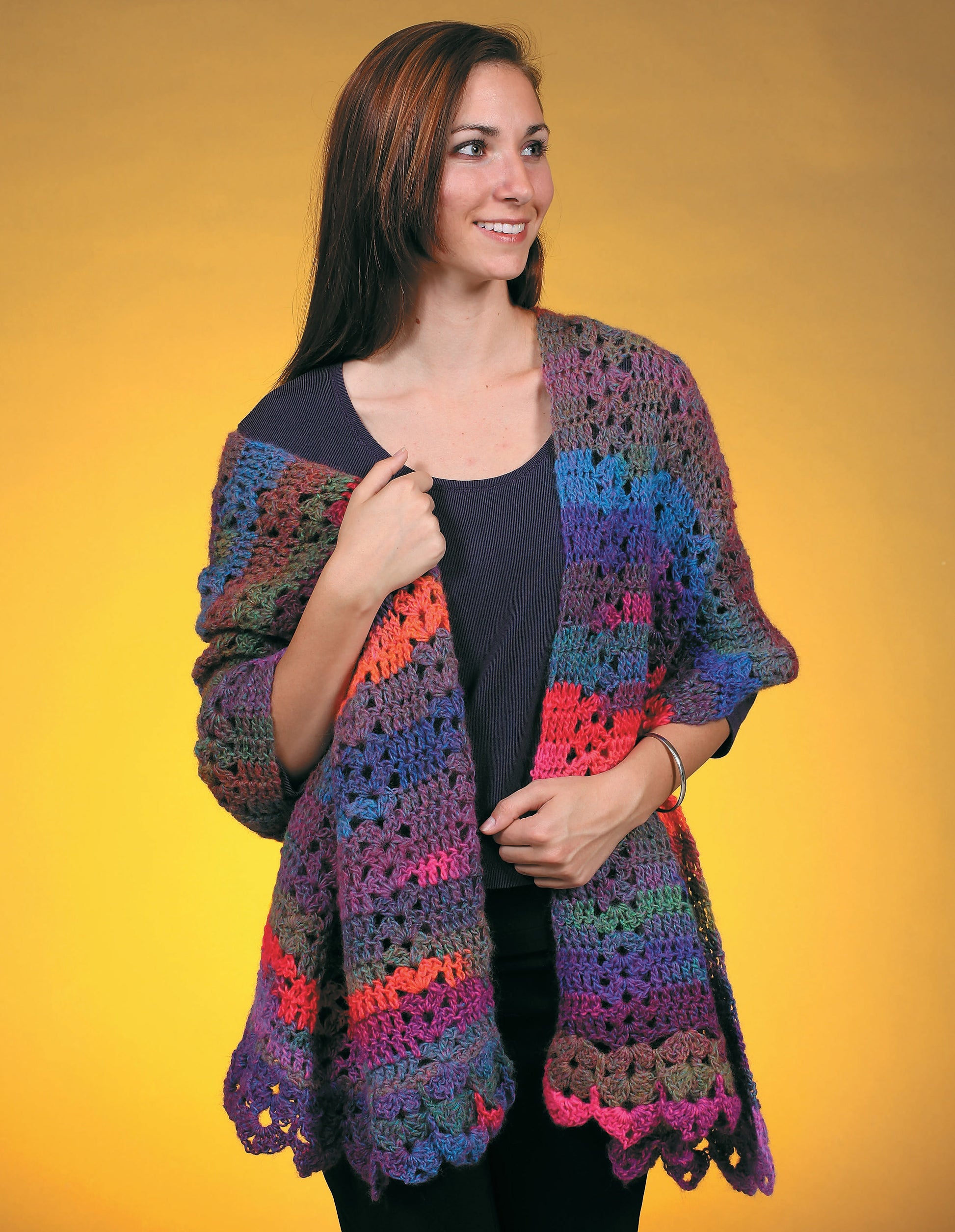 A woman with long brown hair smiles while wearing a colorful crocheted shawl from Leisure Arts' "Make in a Weekend Shawls," over a dark shirt, standing against a yellow background. Perfect for those seeking crochet shawl patterns.