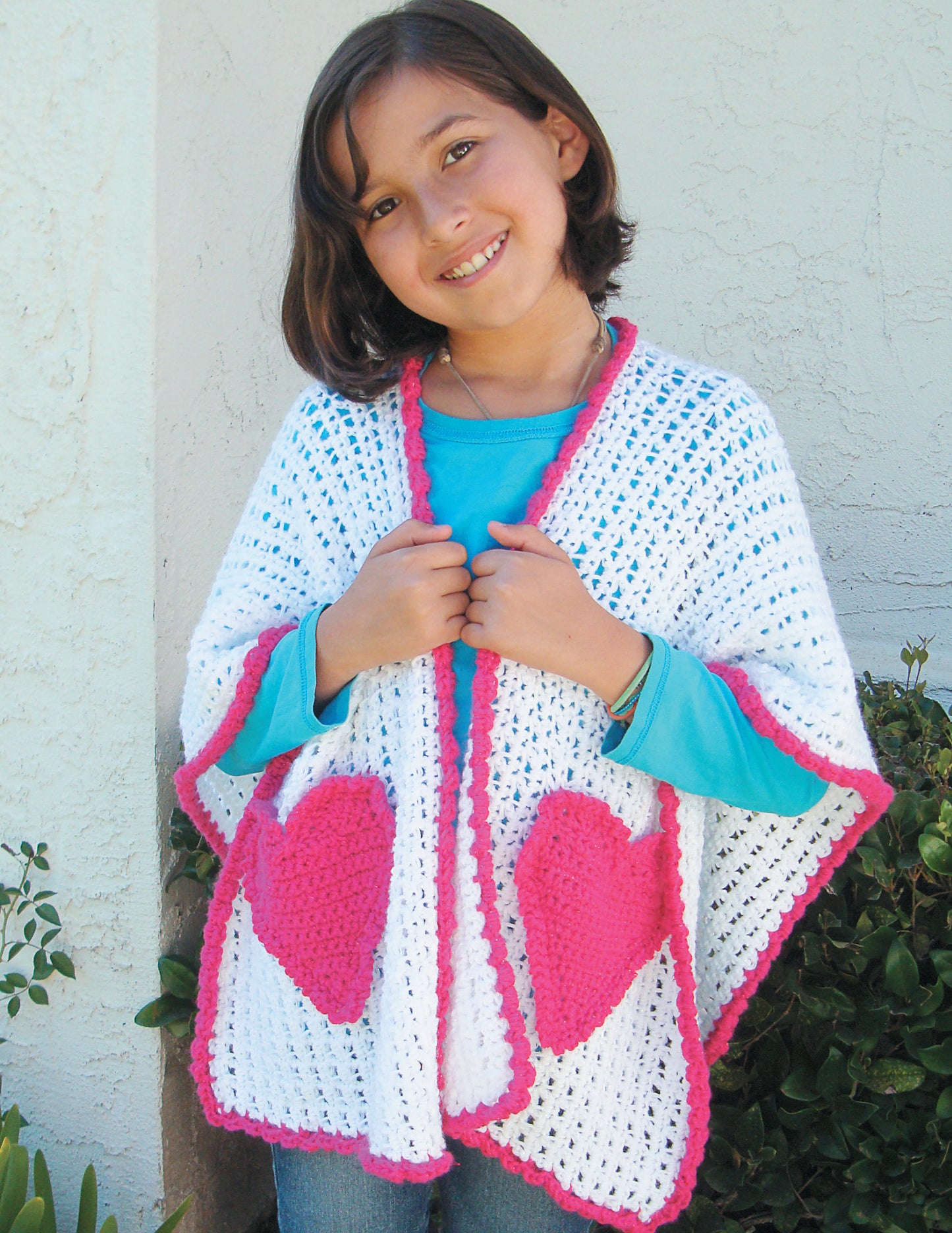 Smiling young girl with brown hair stands outdoors, modeling a white crocheted shawl with pink trim and hearts from "Make in a Weekend Shawls" by Leisure Arts. Light wall and greenery make the perfect backdrop for this fun project.