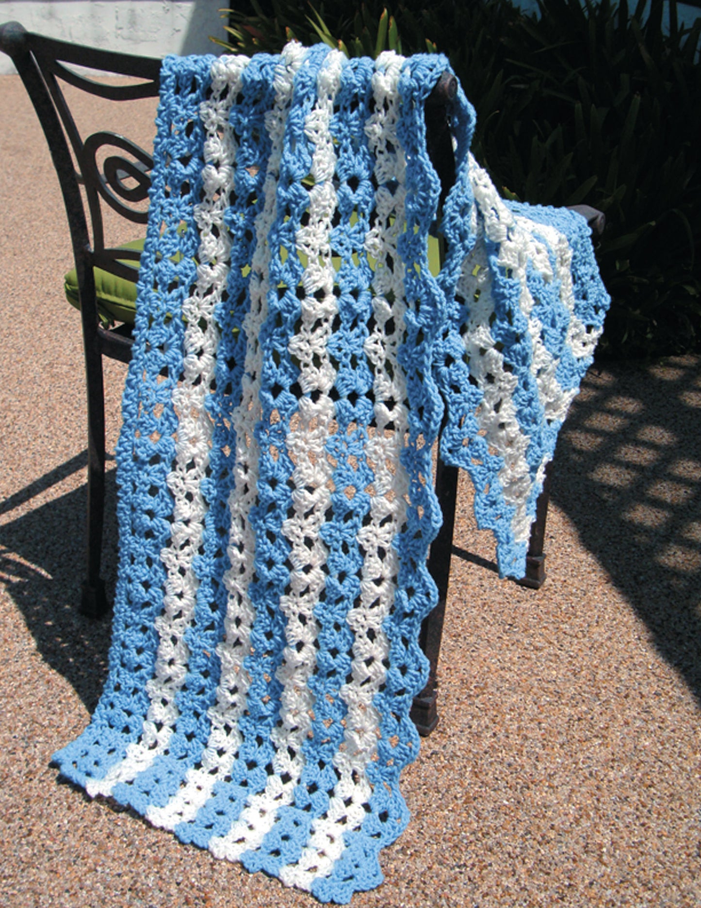 The "Make in a Weekend Shawls" by Leisure Arts, featuring blue and white scalloped stripes, is draped over a metal chair on a pebble patio with greenery in the background—ideal for crochet fans of all ages.
